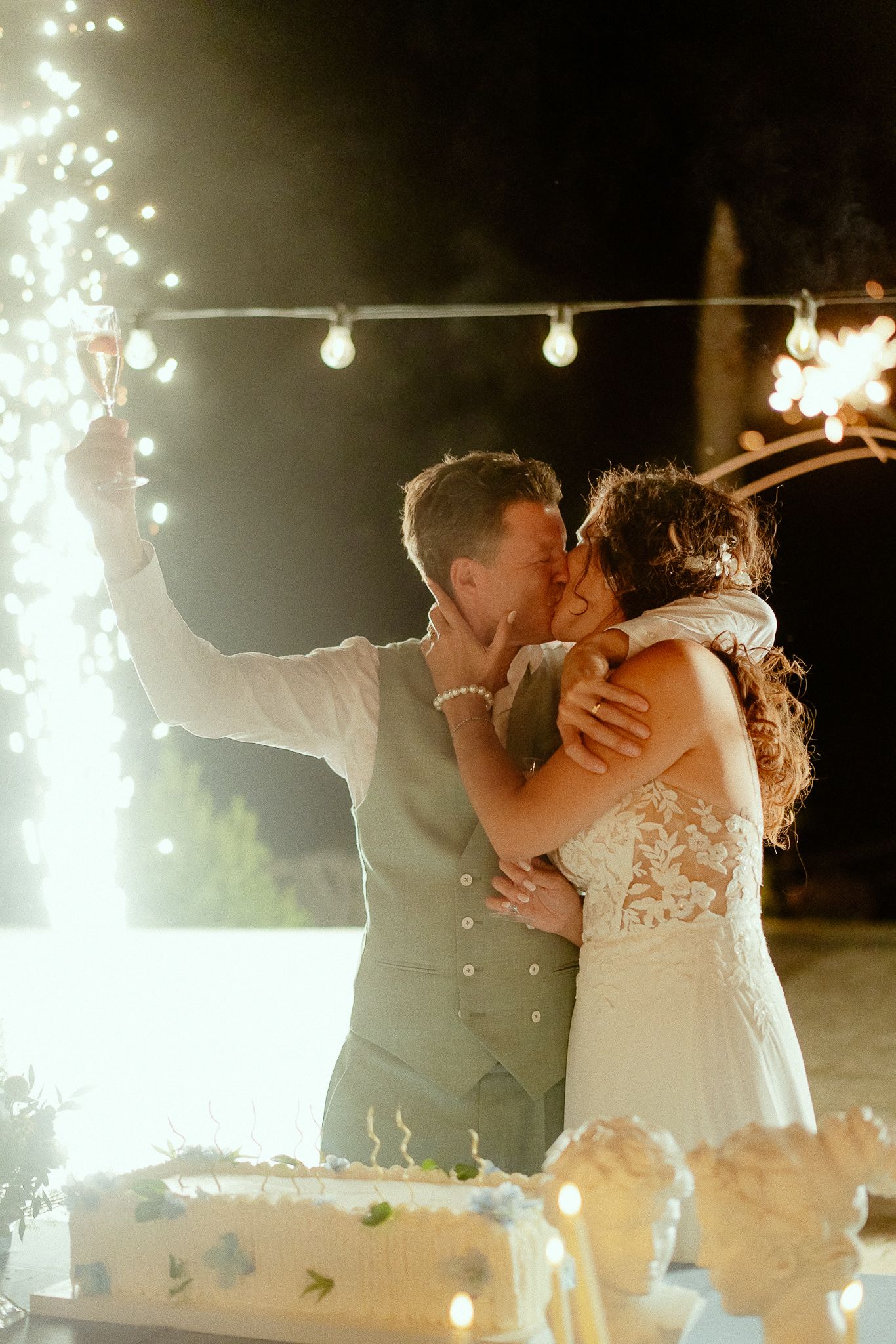 Bride and groom sharing a romantic kiss while holding sparklers beside their wedding cake at an outdoor evening reception.
