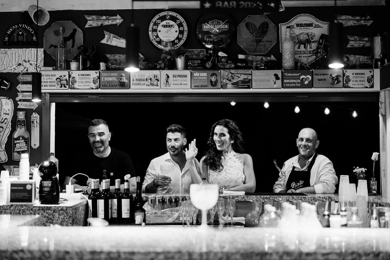 Four smiling bartenders pose behind a bar counter in a lively pub filled with signs and bottles.