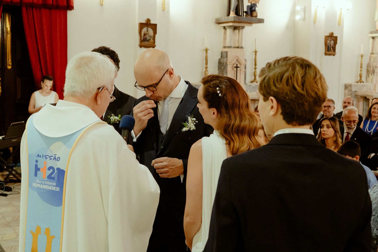 Groom receiving blessing from priest during wedding ceremony in a church, with bride and guests watching.