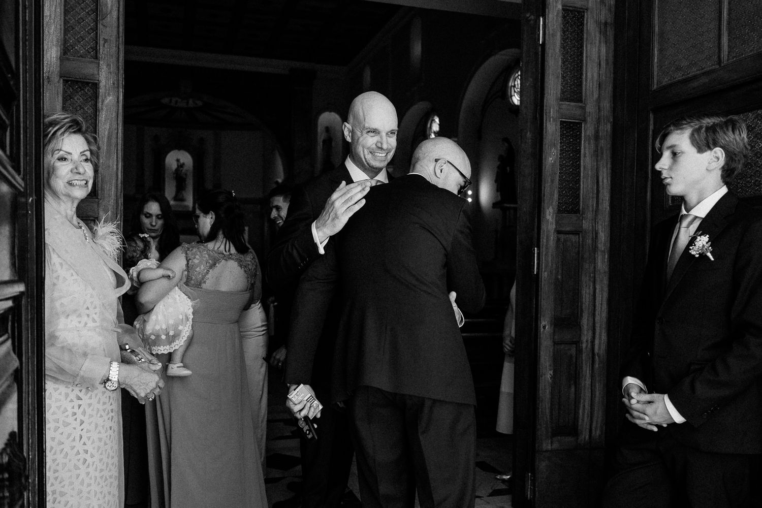 Black and white wedding photo of guests embracing near church doors during a formal ceremony celebration.