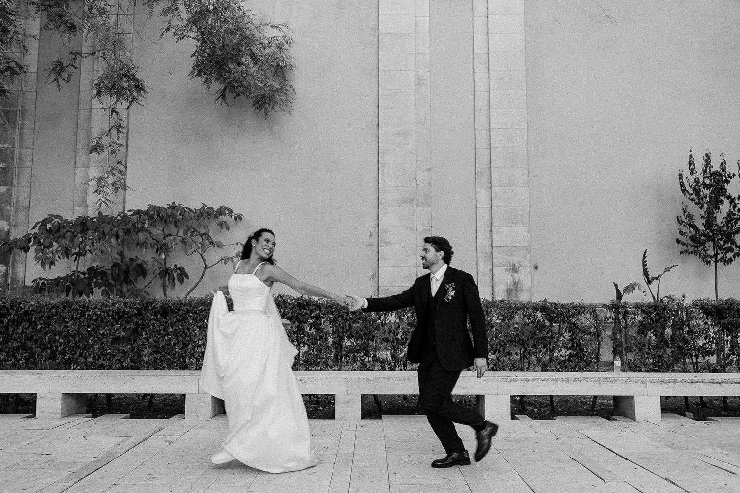 Black and white photo of a bride and groom holding hands, walking outdoors near hedges and trees on their wedding day.
