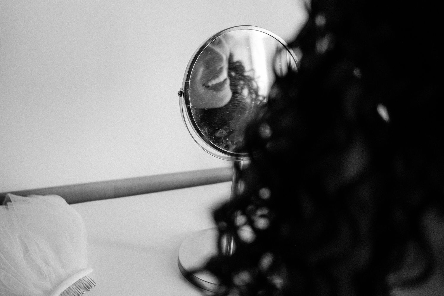 Black and white photo of curly hair beside a round mirror reflecting a blurred image on a white surface.
