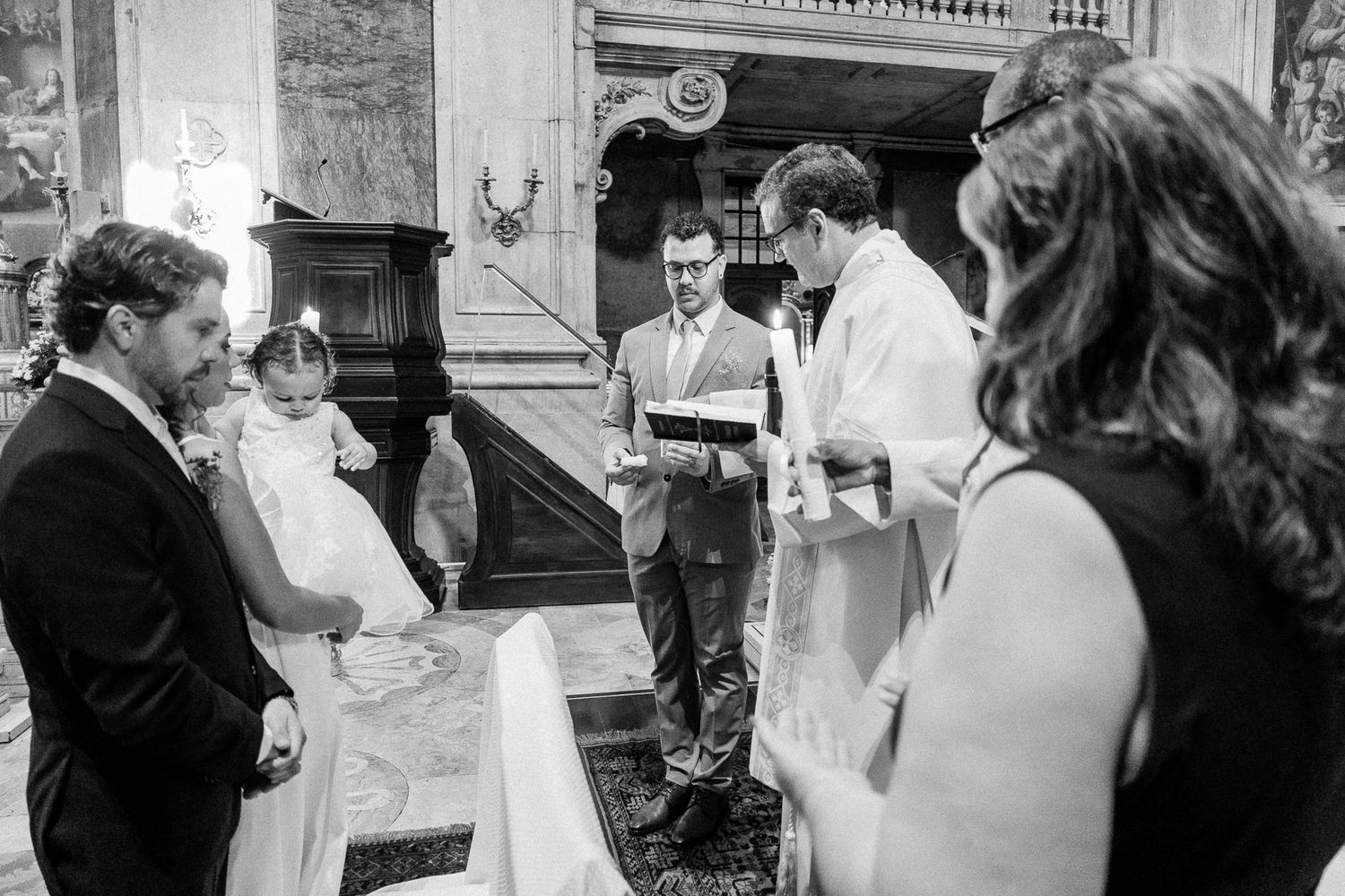 Black and white photo of a baptism ceremony in a church with a priest, parents, and baby in white gown.