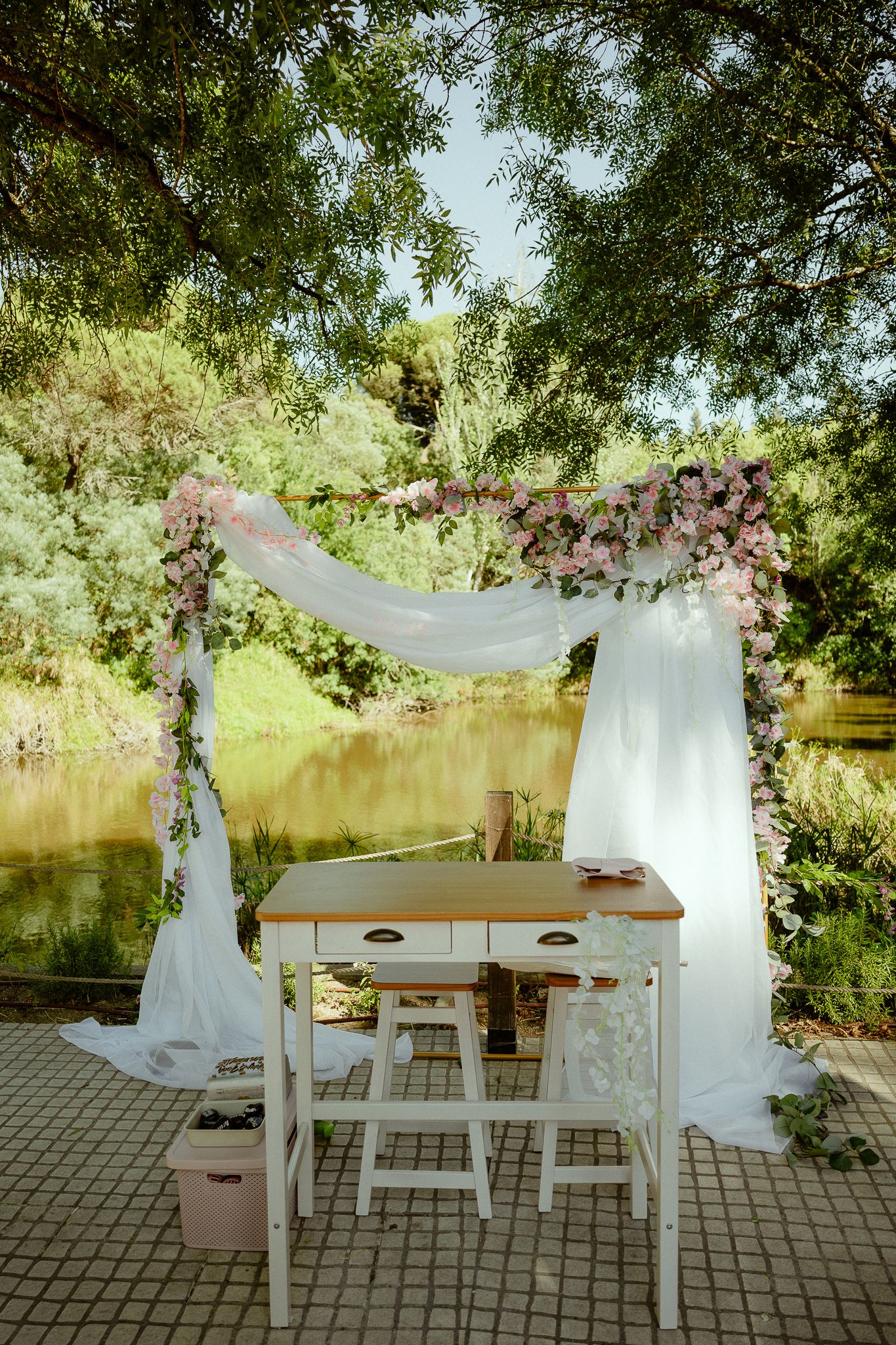 Elegant outdoor wedding arch with white draping and pink flowers beside a serene lake surrounded by lush green trees.
