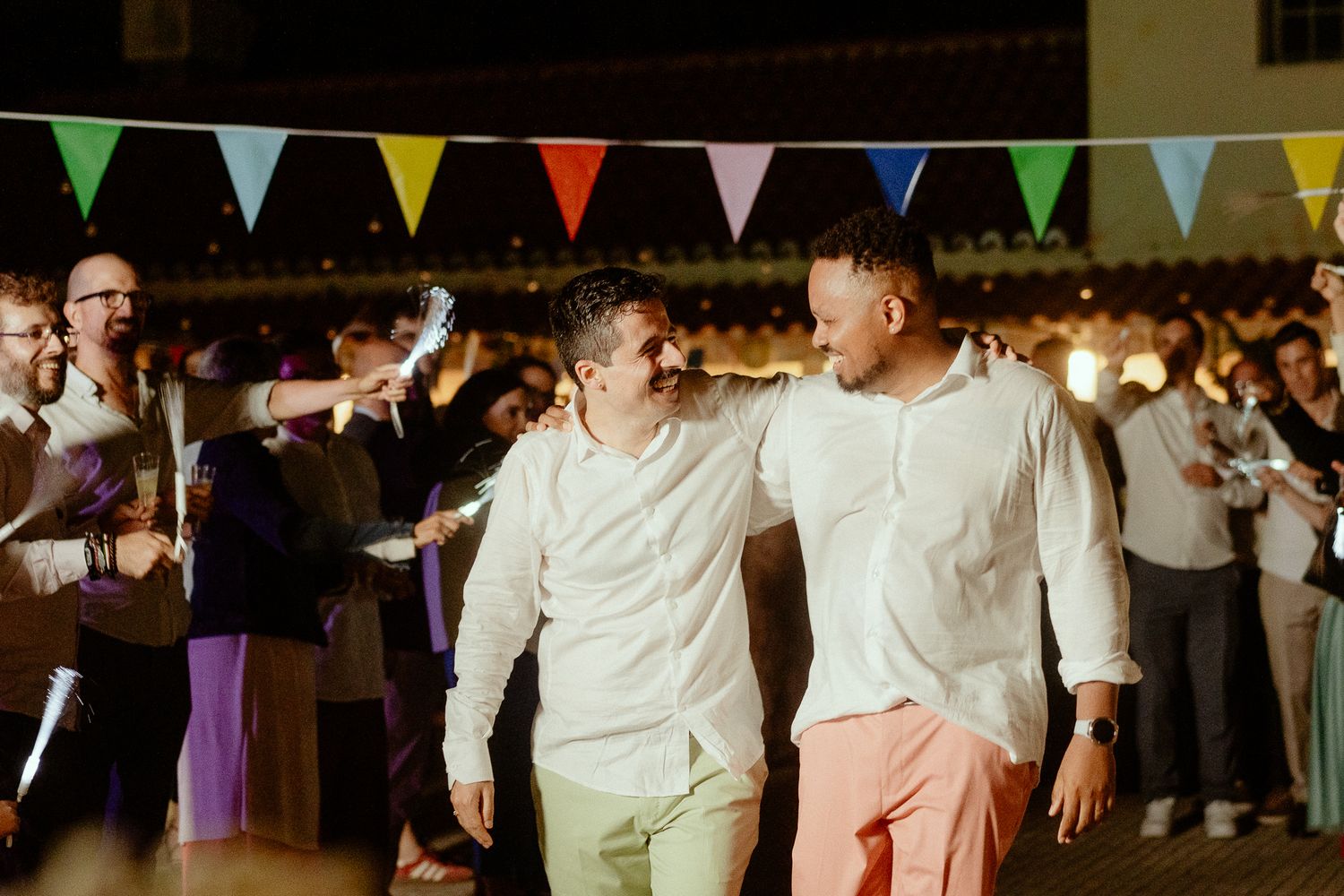 Two grooms in white shirts celebrate at their wedding reception under colorful bunting lights.