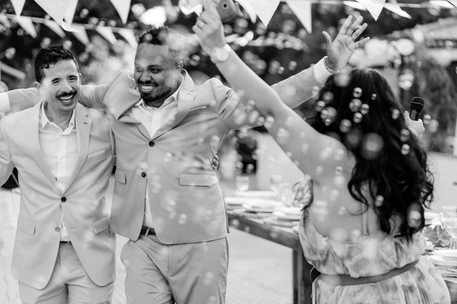 Black and white wedding photo of two grooms celebrating with a guest amid bubbles at an outdoor reception.