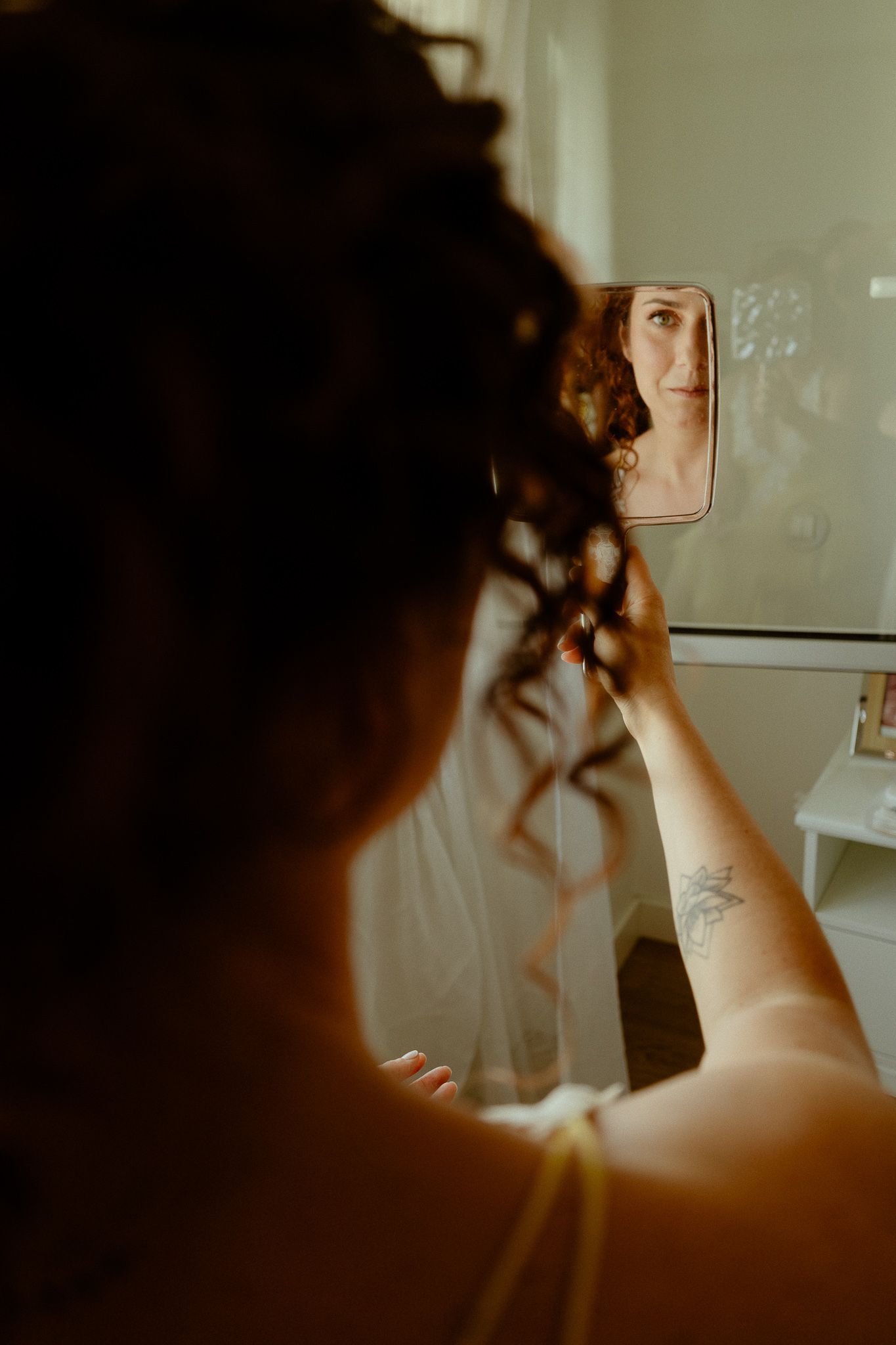 Bride with curly hair and tattoo holds a small mirror reflecting her smiling face while getting ready.