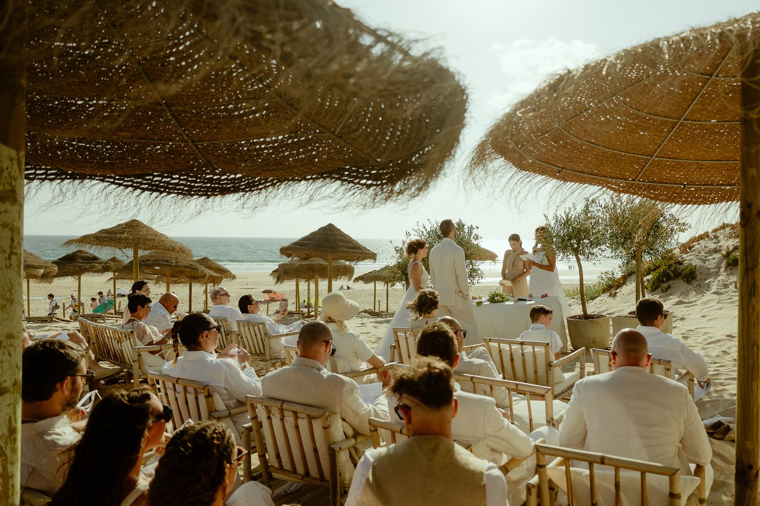 Guests in white attire attend a beach wedding ceremony under straw umbrellas with ocean views.
