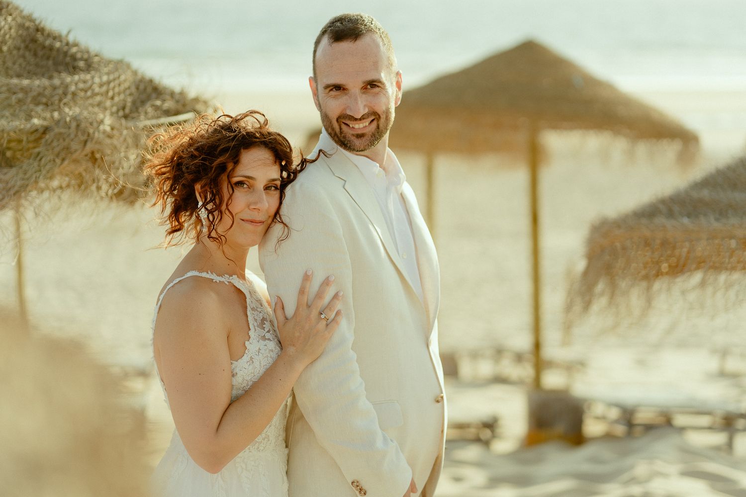 Couple in wedding attire posing on a sunny beach with straw umbrellas in the background.