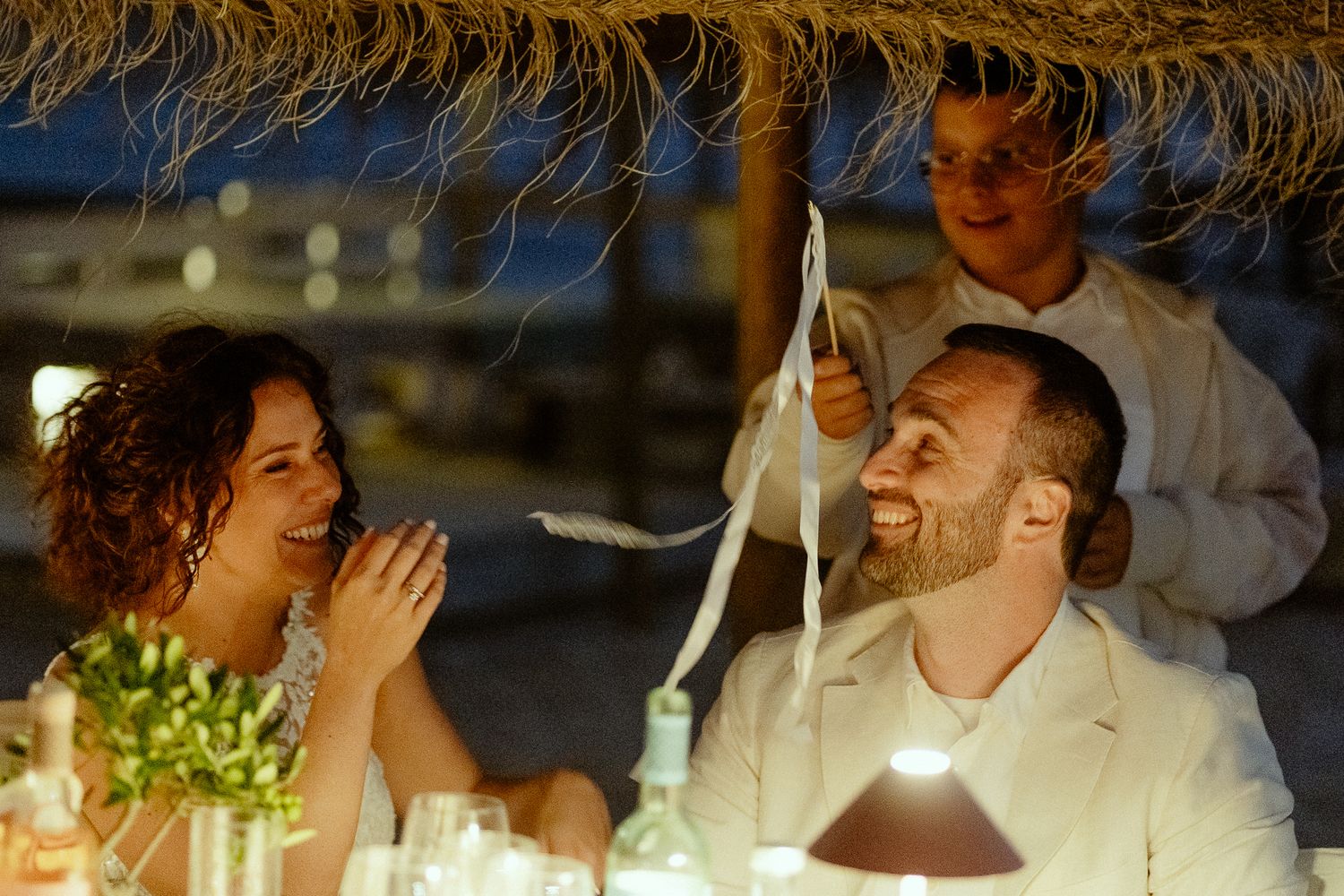 Couple laughing joyfully at outdoor wedding reception dinner under thatched roof at dusk.