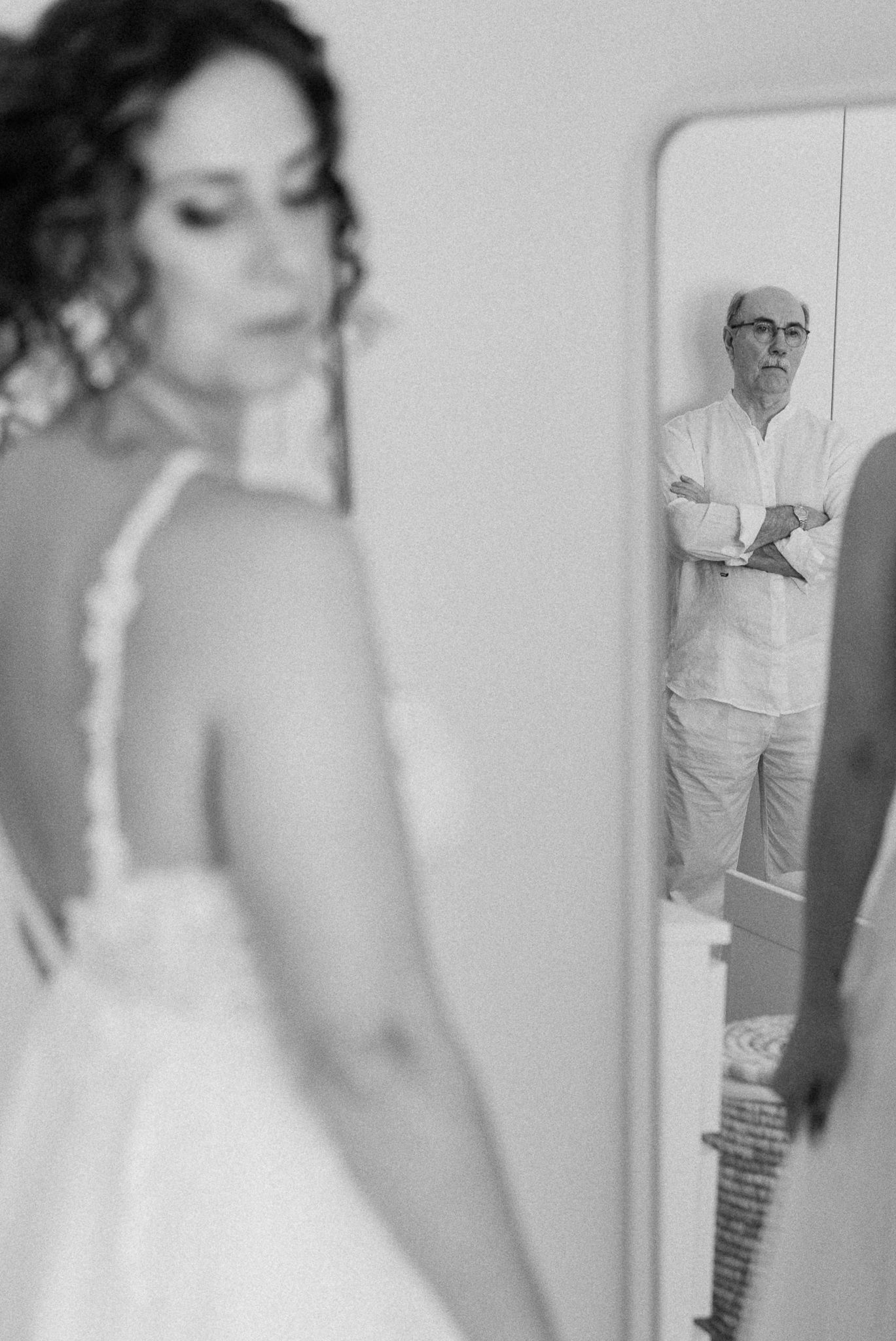 Black and white photo of bride in wedding dress with father's reflection visible in mirror behind her.