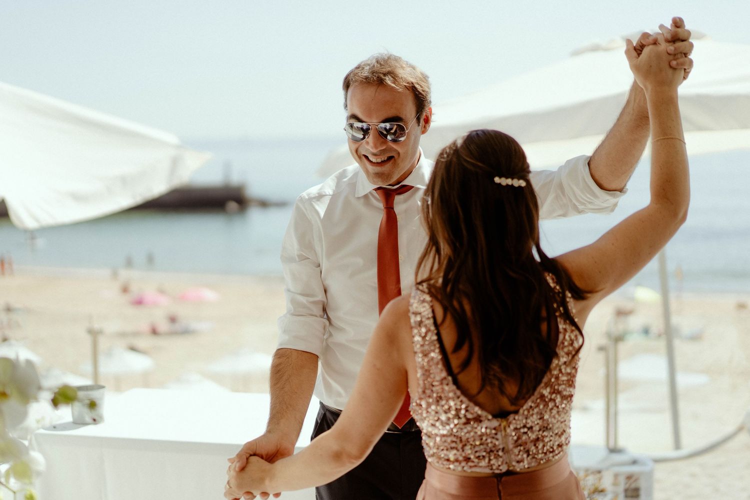 A couple dances together on a sunny outdoor terrace overlooking a beach with white umbrellas in the background.