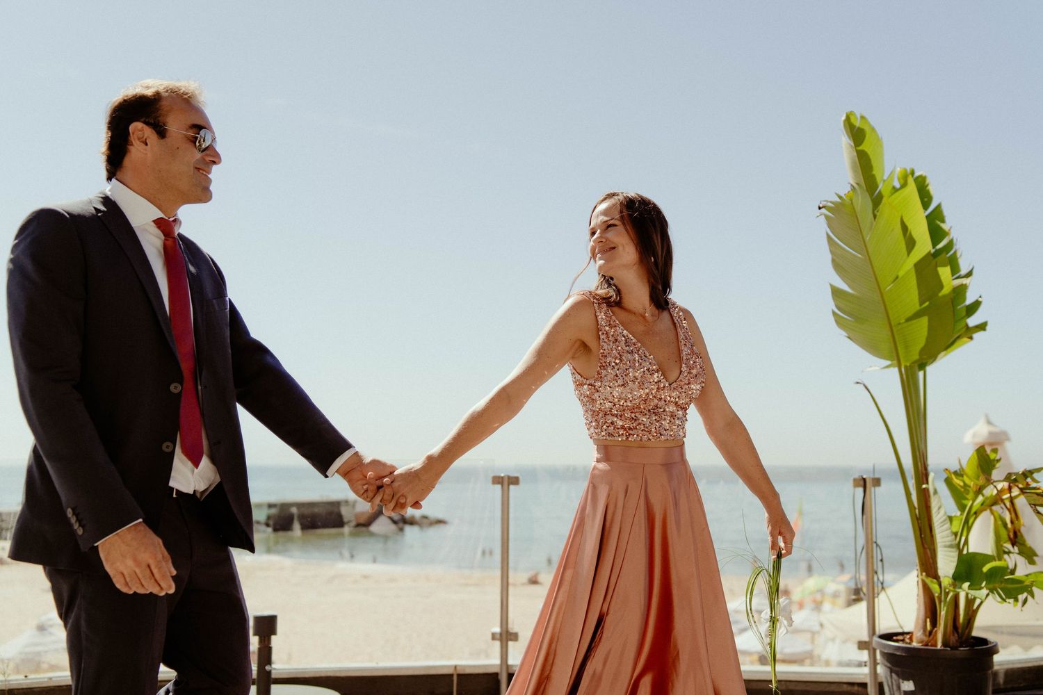A couple holds hands and dances together on a sunny beach with palm trees and ocean views in the background.