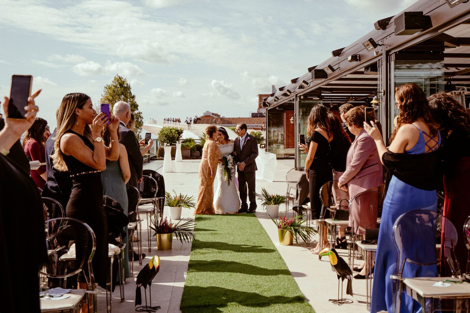 Outdoor wedding ceremony with guests standing along green aisle runner.