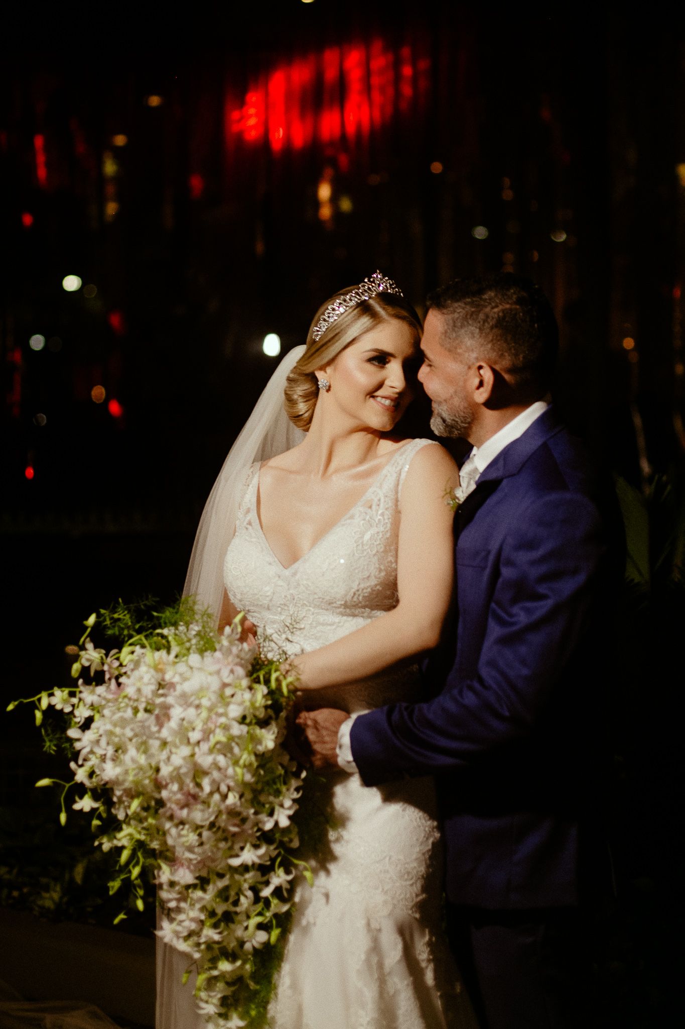 A romantic nighttime wedding portrait with a lace-dressed bride holding a cascading white bouquet against dark scenery.