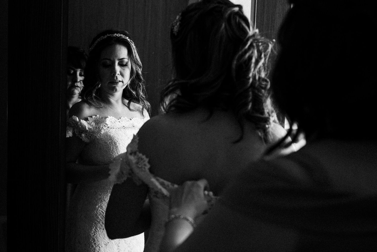 A person in a white lace wedding dress looks in a mirror in an intimate black and white photo.