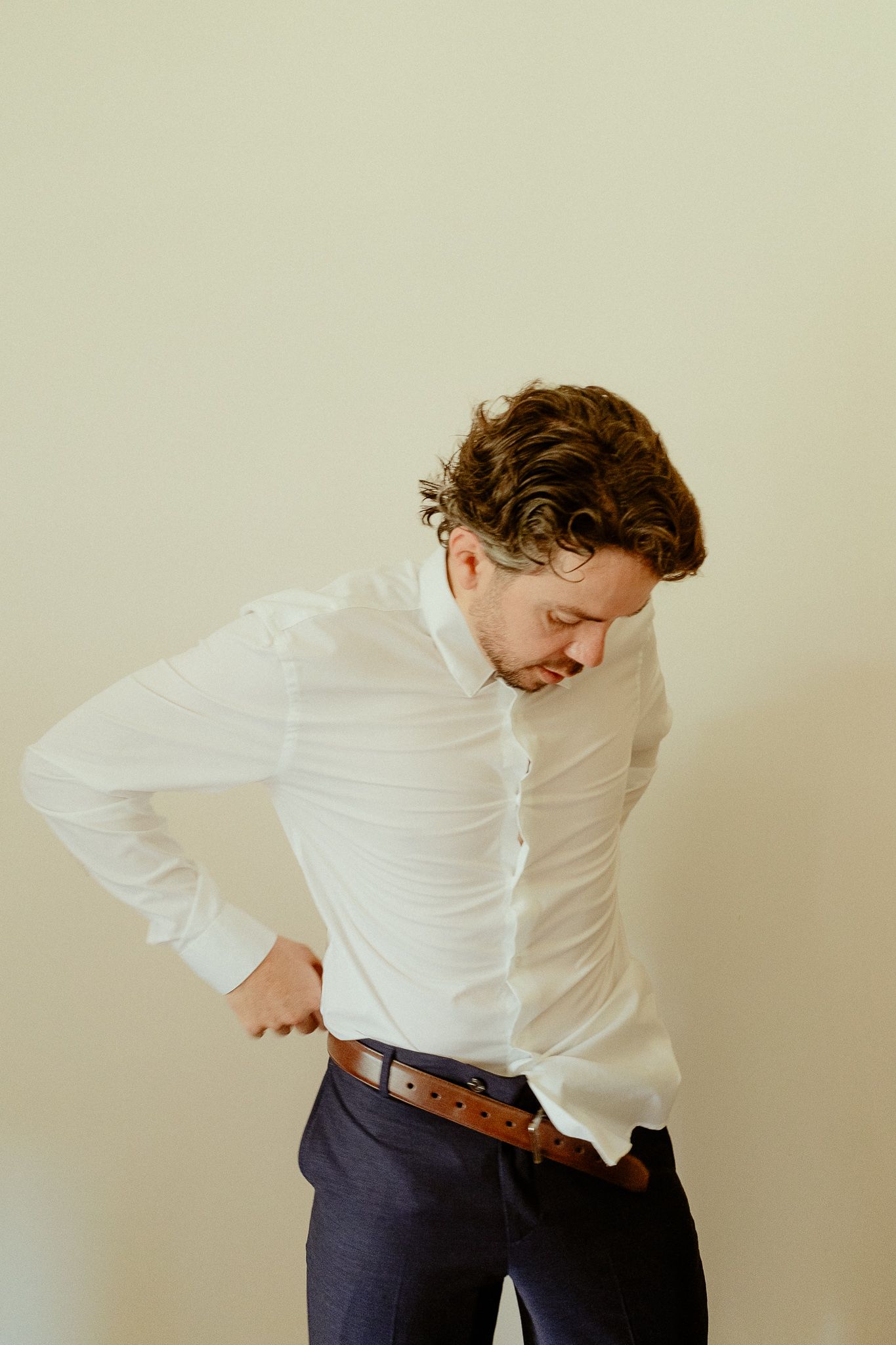 A man in a white dress shirt and navy pants adjusting his brown leather belt against a neutral background.
