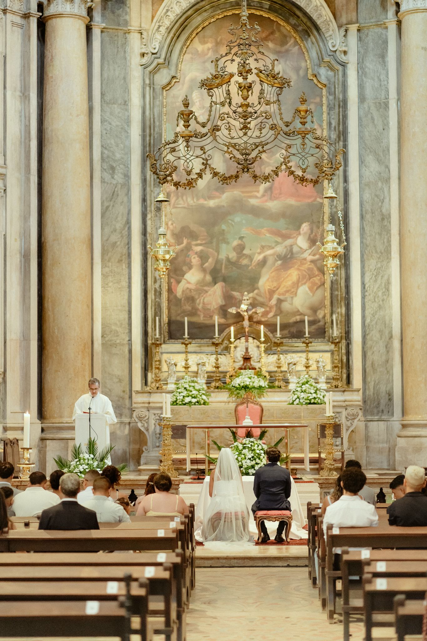 Elegant church wedding ceremony with bride and groom kneeling at ornate baroque altar with gold details and classical painting.