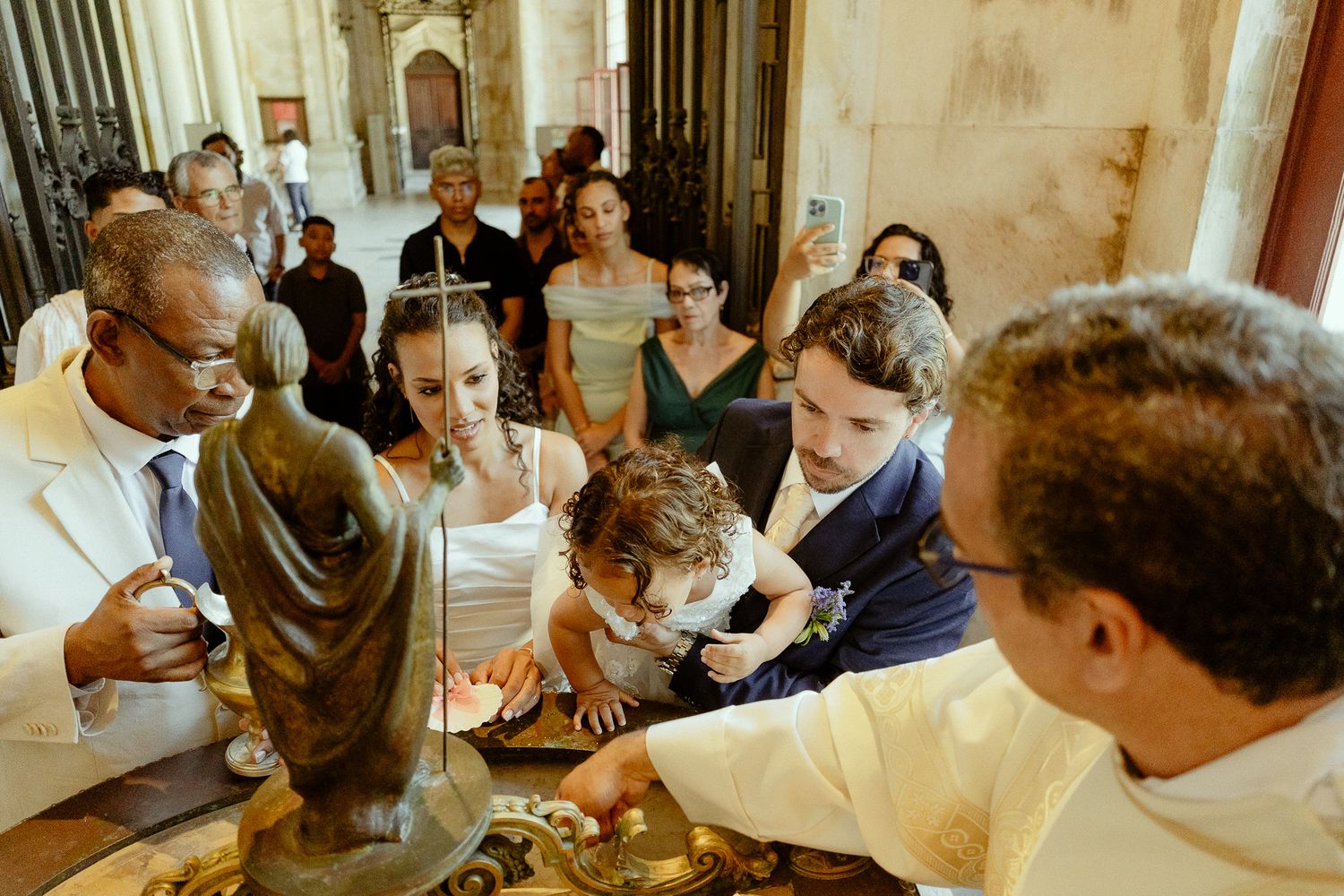 A Catholic baptism ceremony with a priest, parents, and child gathered around a baptismal font in a church.