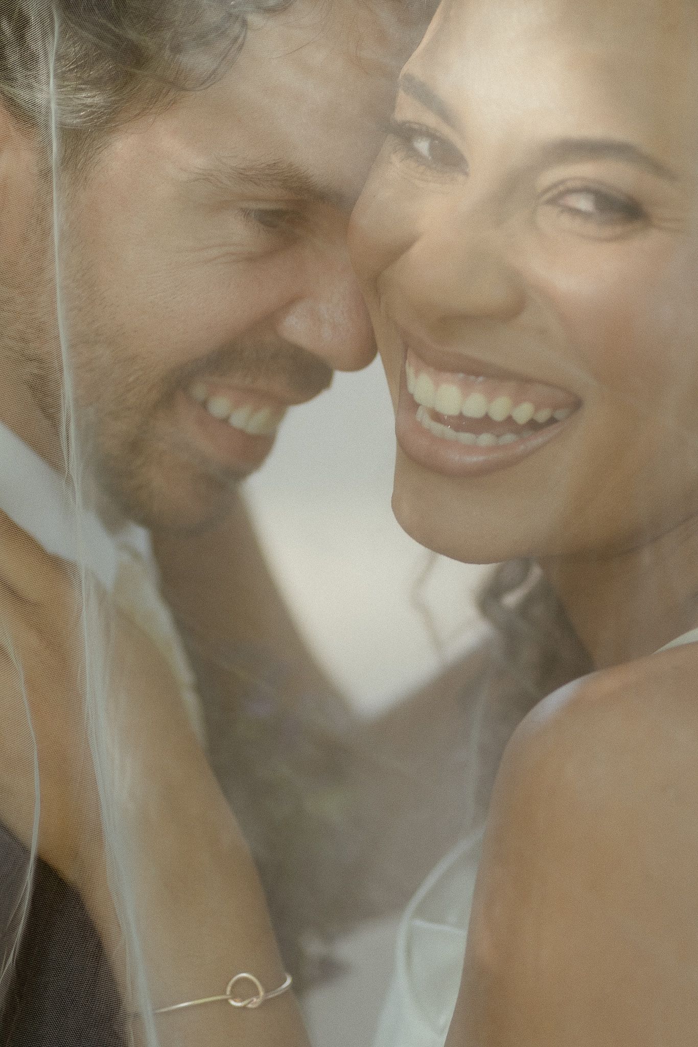 Happy bride and groom laughing together under a wedding veil on their special day.