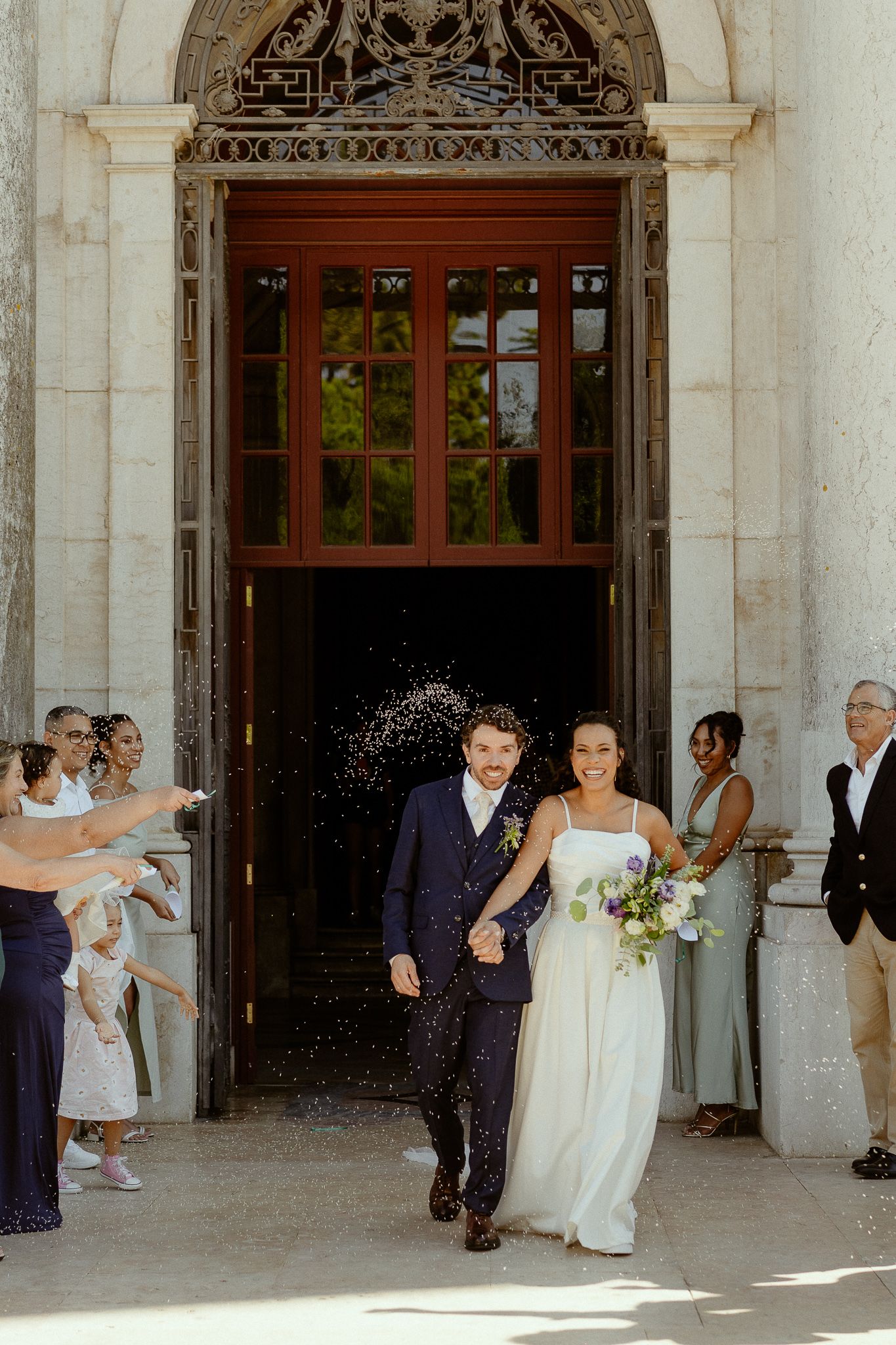 Newlywed couple exits church doors as guests toss confetti, bride holding bouquet, groom in navy suit.