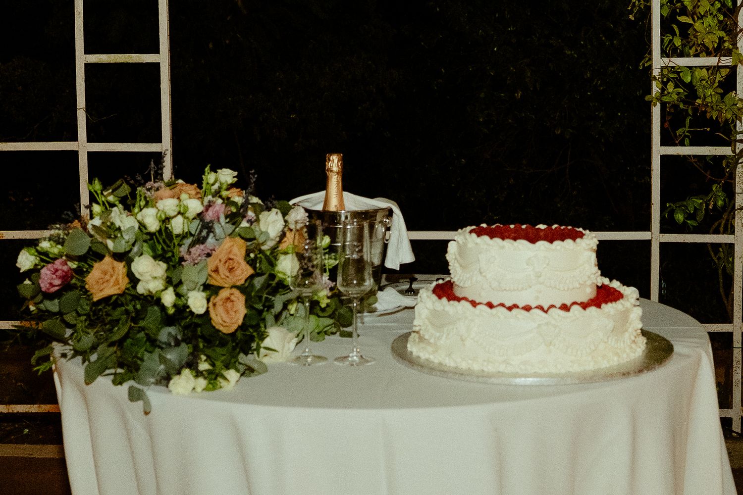 Two-tier white wedding cake with red berries beside a floral bouquet and champagne bottle on a draped table.