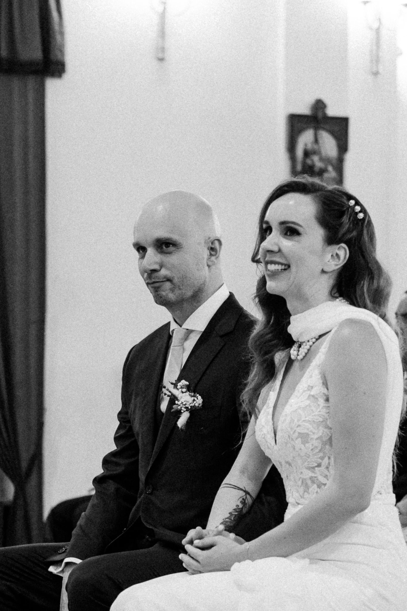 Black and white photo of a smiling bride in lace gown and groom in suit with boutonniere seated at wedding ceremony.