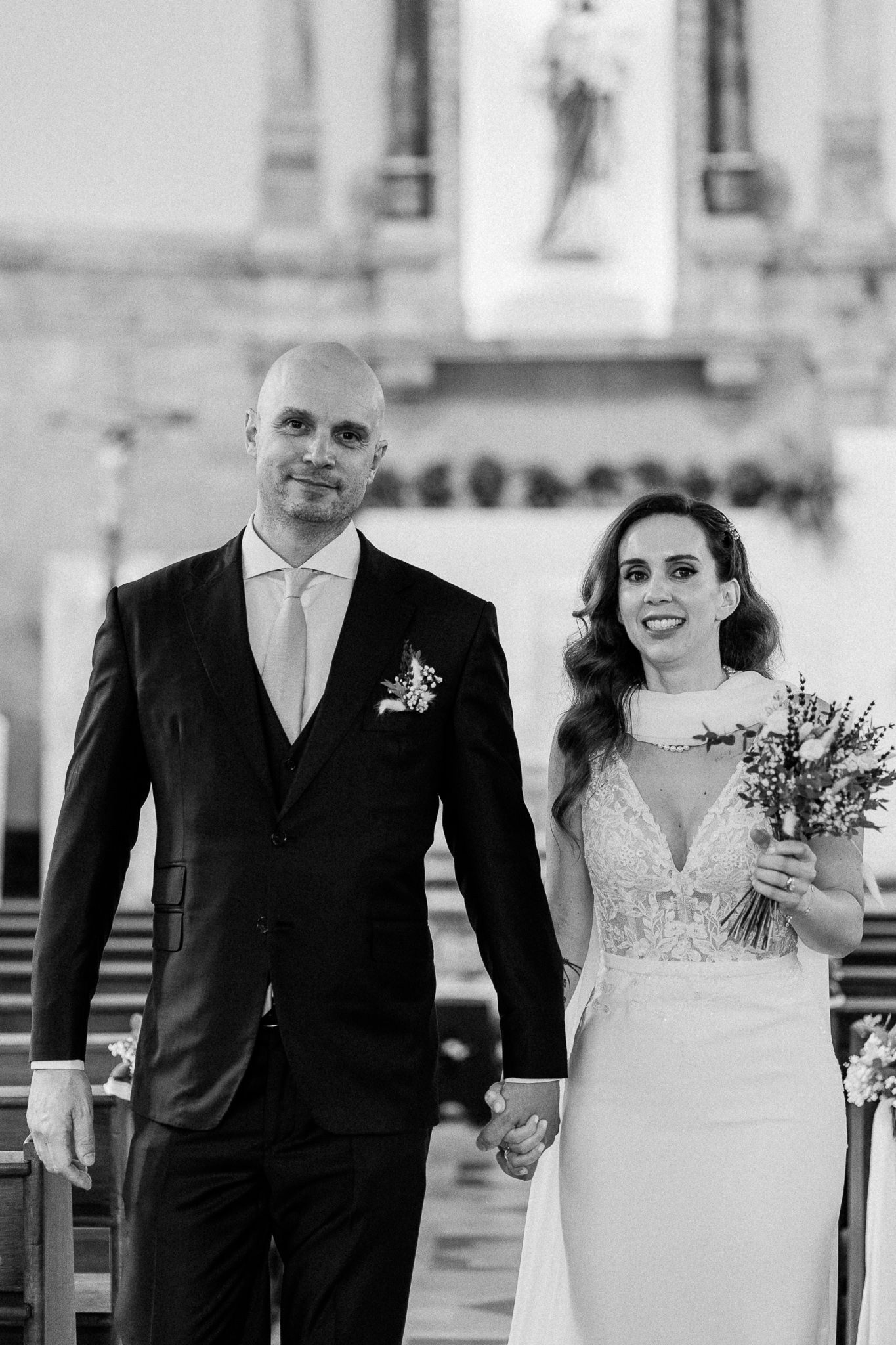 Black and white photo of a smiling bride and groom holding hands at the altar of a church on their wedding day.