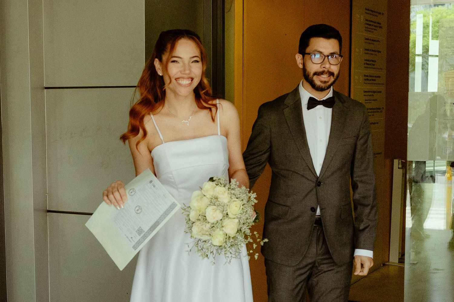 Smiling bride in white dress holding bouquet walks with groom in dark suit and bow tie after wedding ceremony.