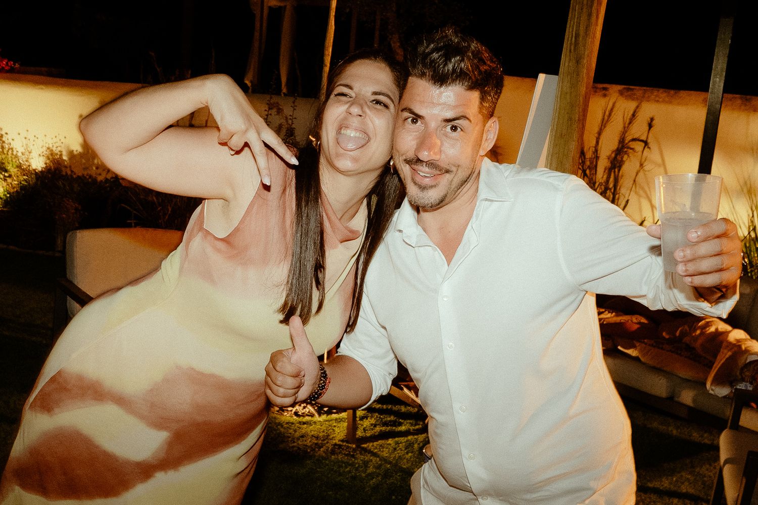Two friends smiling and posing together at an outdoor evening party, holding drinks and giving thumbs up.