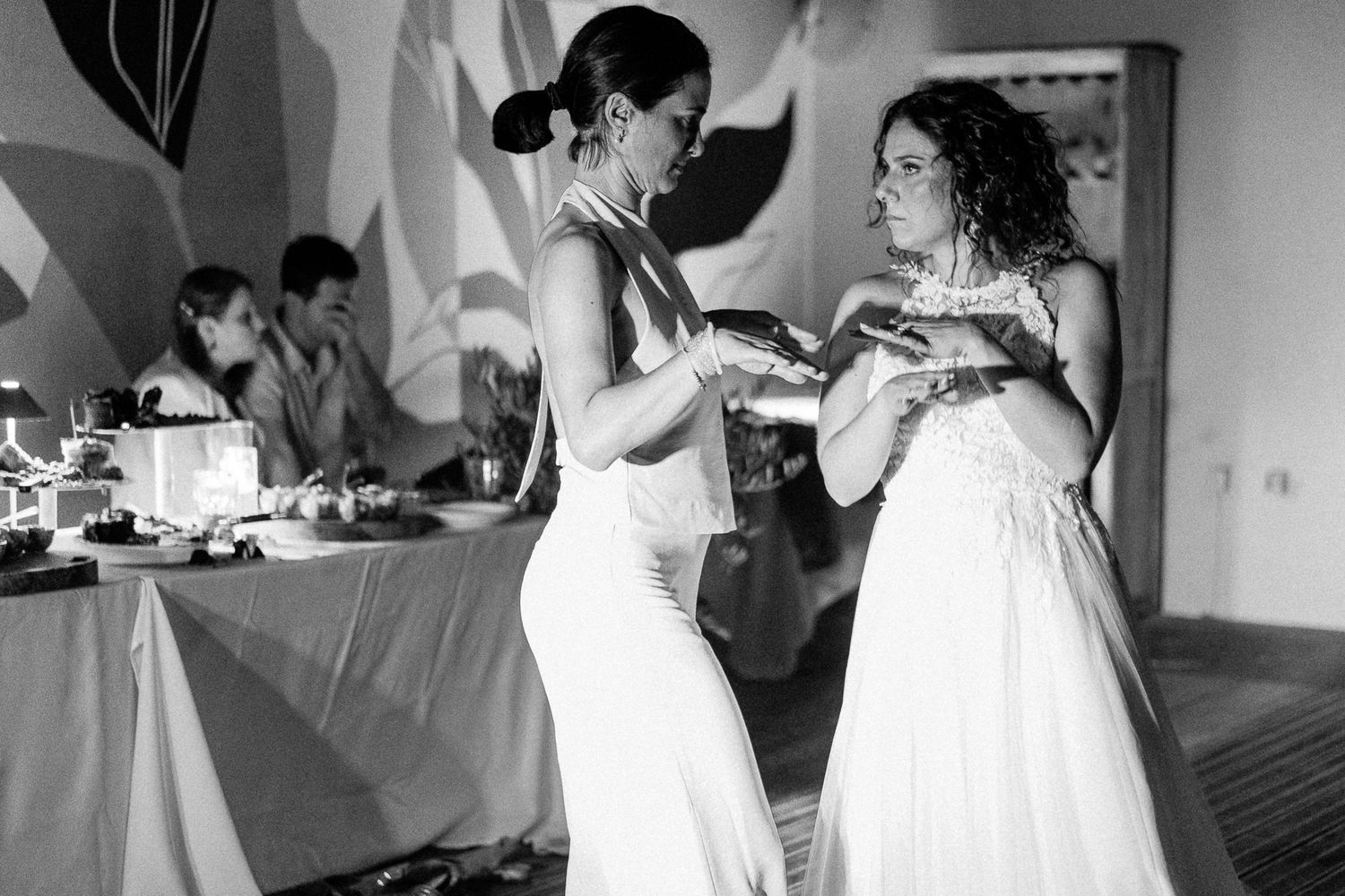 Two women in white dresses share a moment at a wedding reception in this elegant black and white photo.
