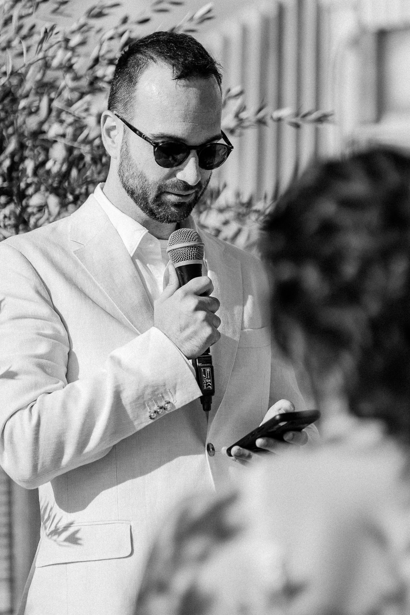 Man in white suit and sunglasses speaking into microphone at outdoor wedding ceremony.