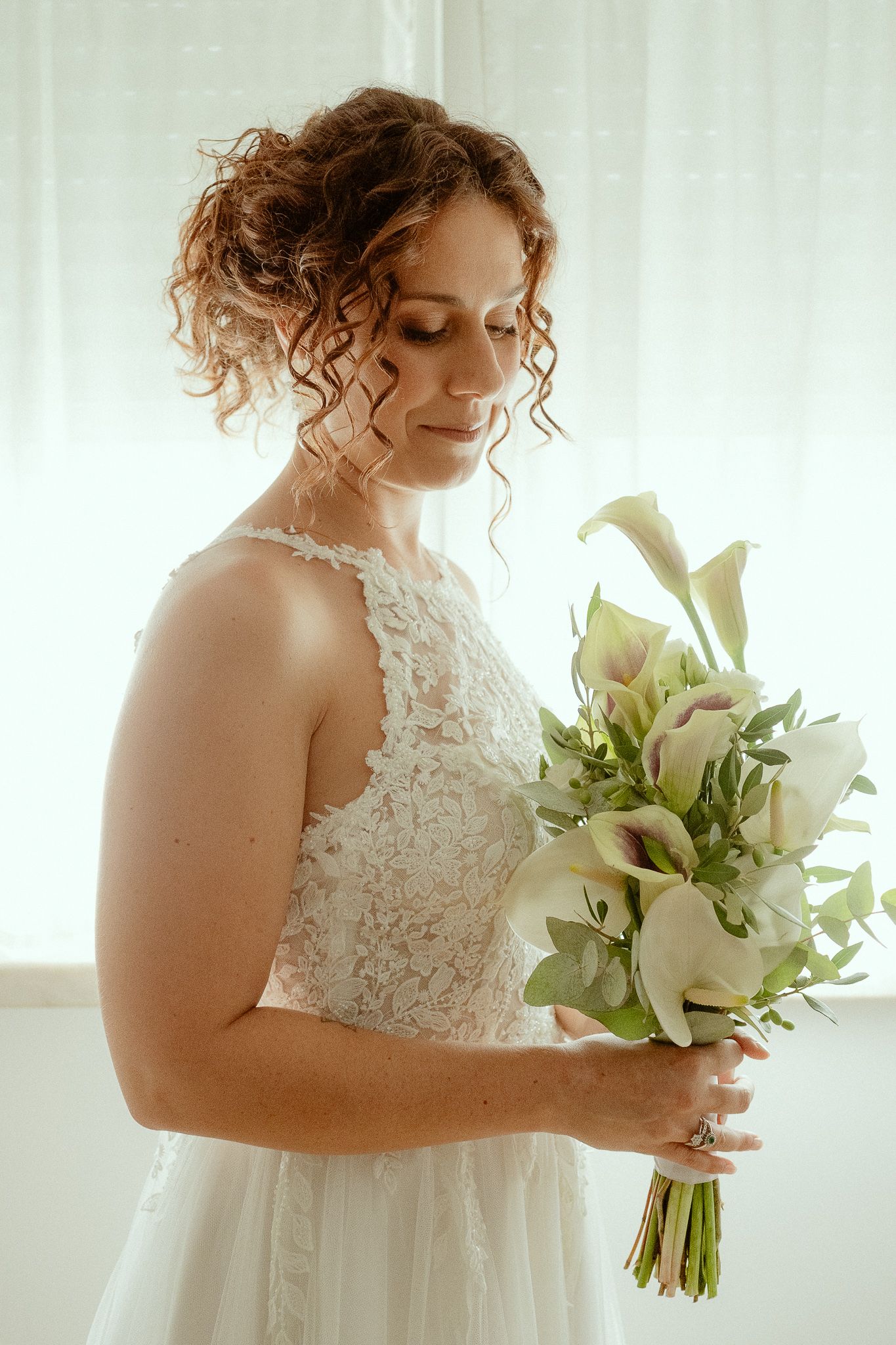 Bride in lace wedding dress holding elegant white calla lily bouquet, standing near bright window with soft natural light.
