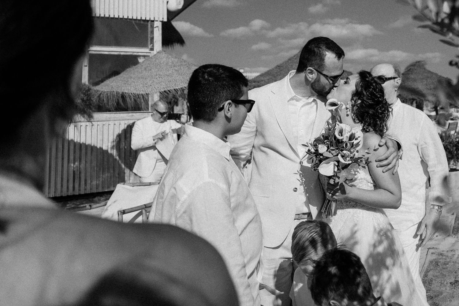 Black and white wedding photo of bride and groom sharing a kiss outdoors, surrounded by guests in white attire.