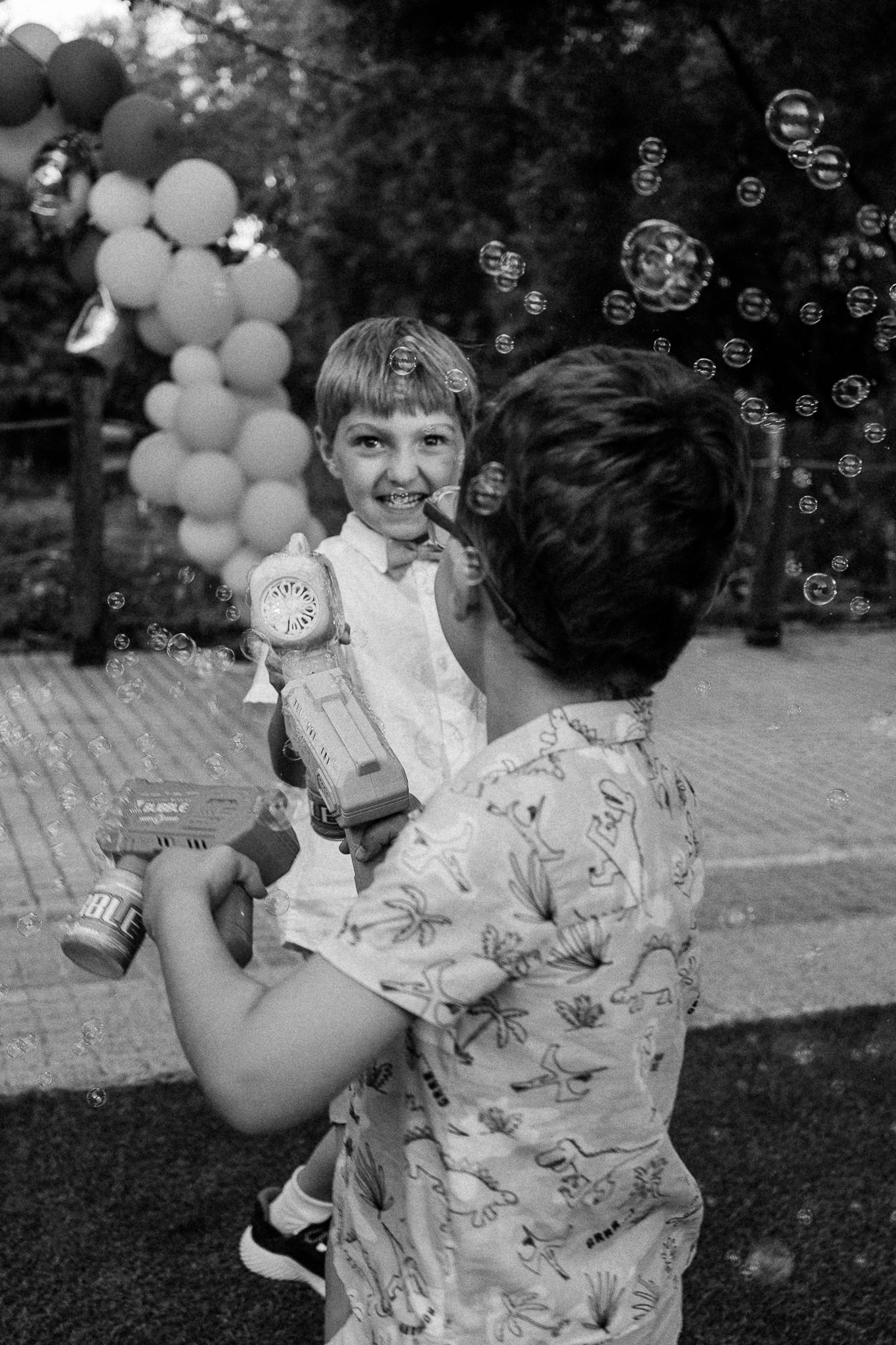 Black and white photo of adult holding smiling child with bubble gun at outdoor birthday party with balloon decorations.