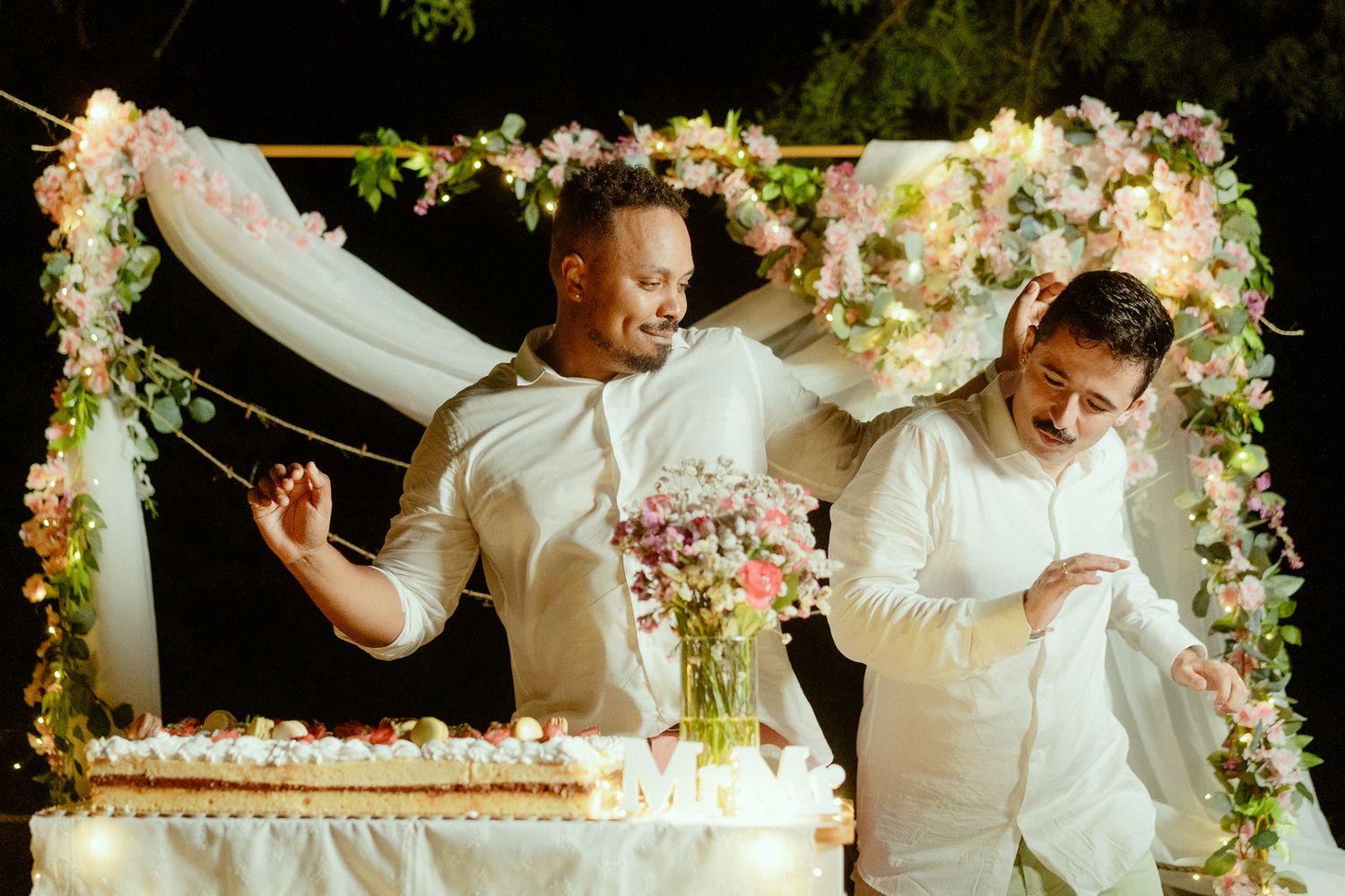Two grooms in white cutting wedding cake under floral arch with pink and white flowers at outdoor night ceremony.
