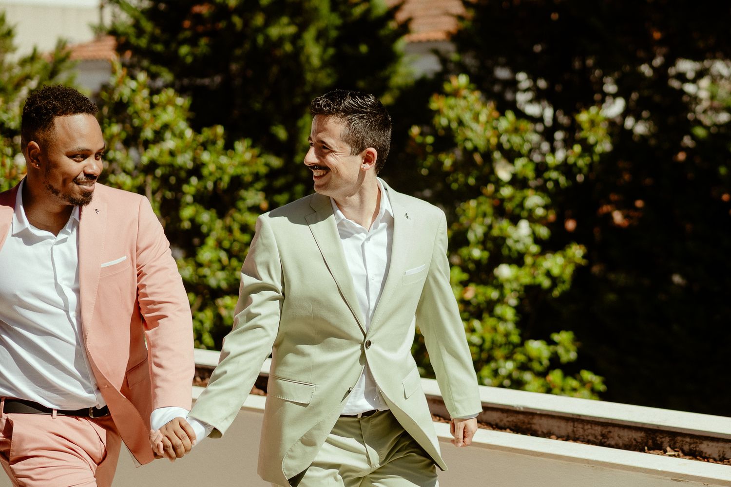 Two men in colorful blazers walk hand in hand outdoors, smiling joyfully on a sunny day.