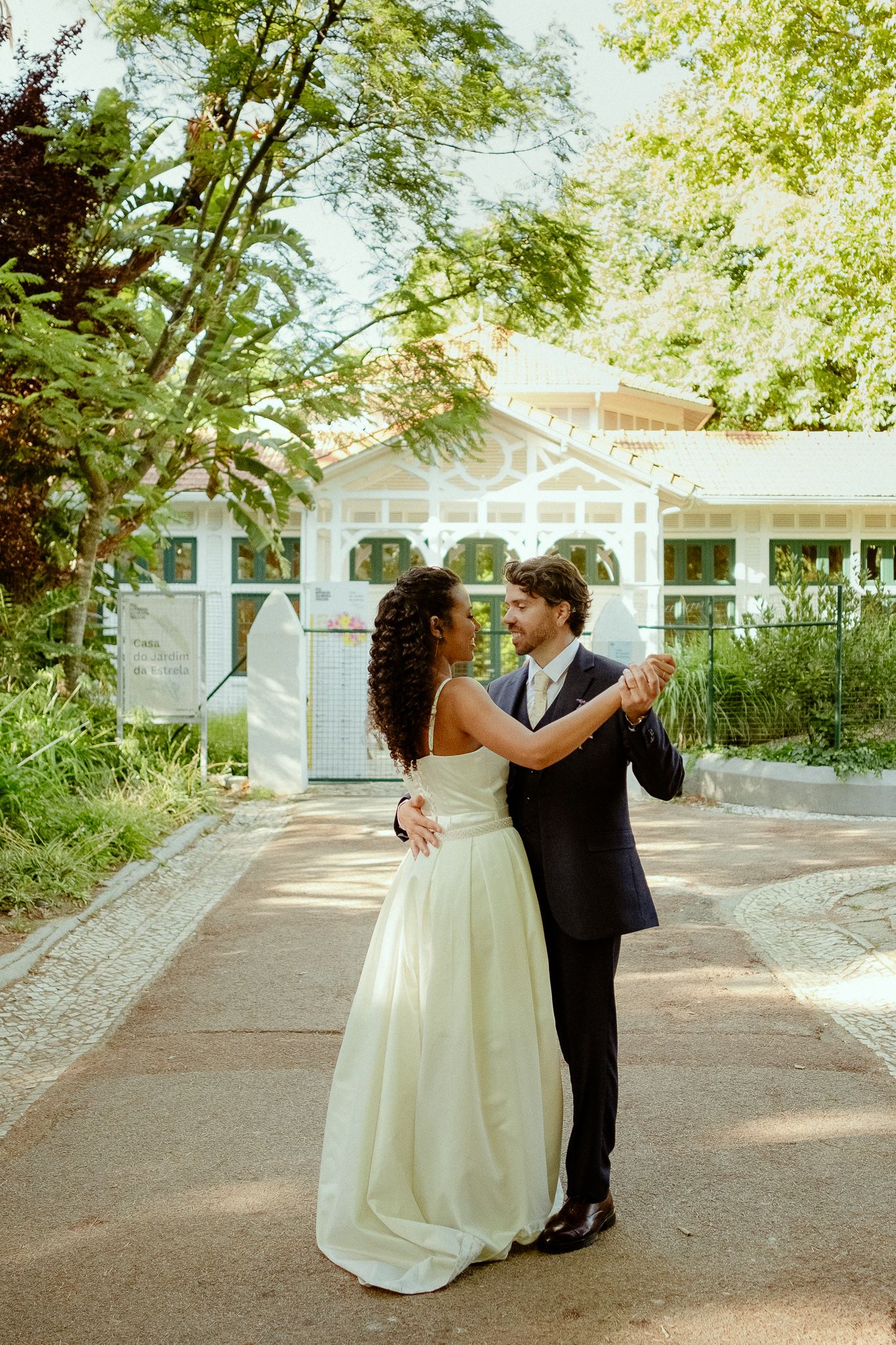 A bride and groom share a first dance outdoors in a lush garden setting, with a white pavilion in the background.