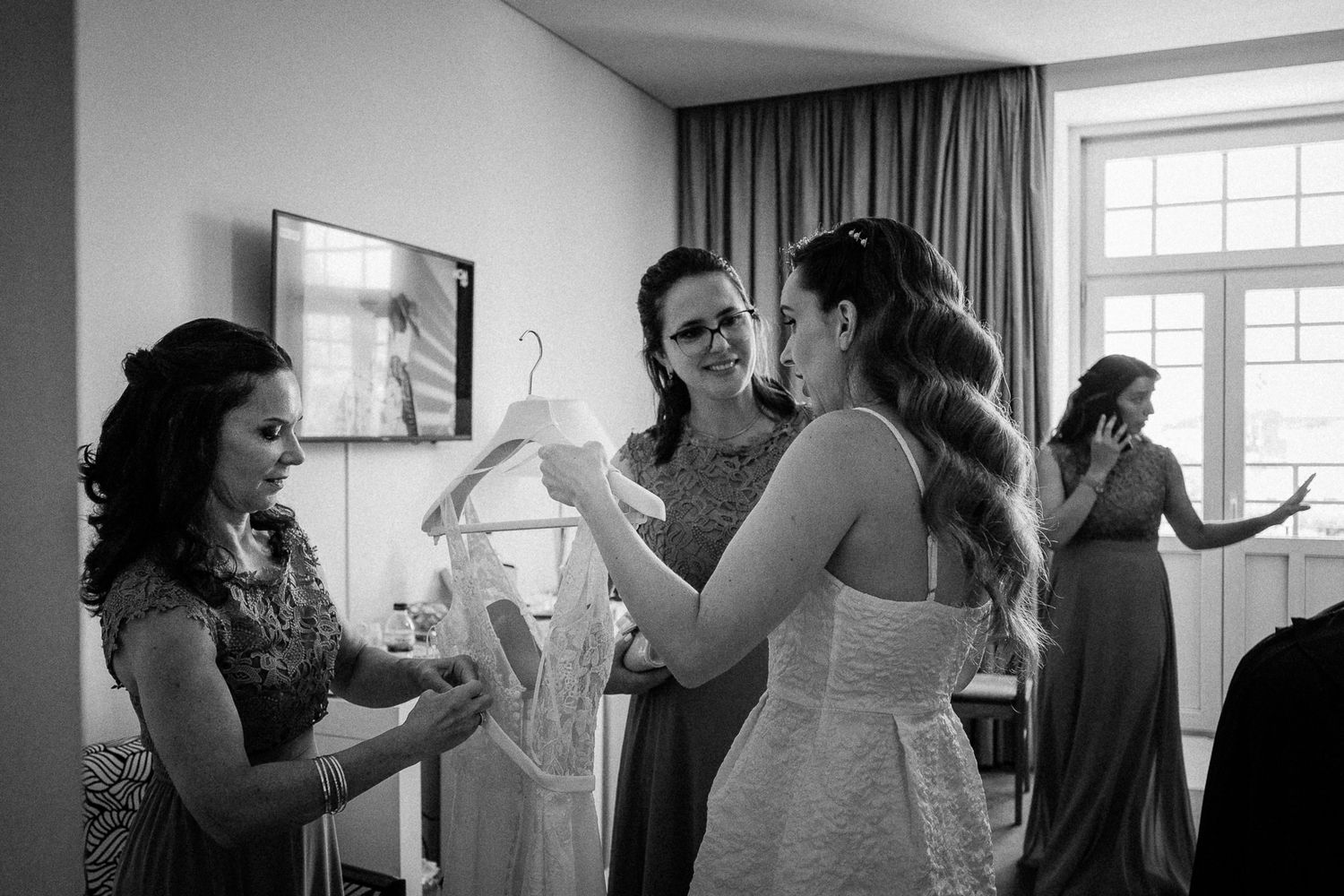 Black and white photo of bridesmaids helping bride with wedding dress in a bright hotel room.