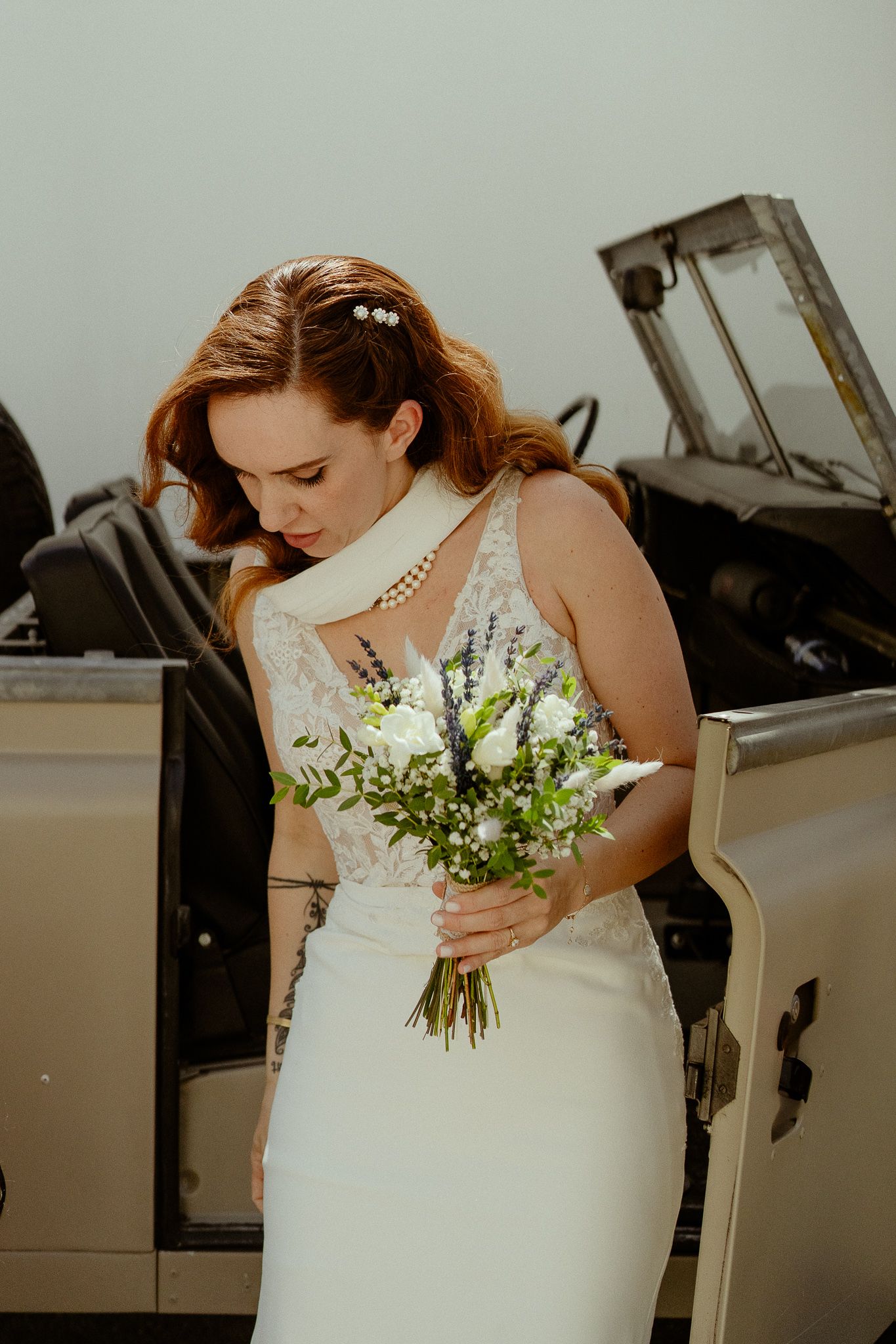 A bride in a white lace gown holds a bouquet of white roses and greenery beside a vintage car.