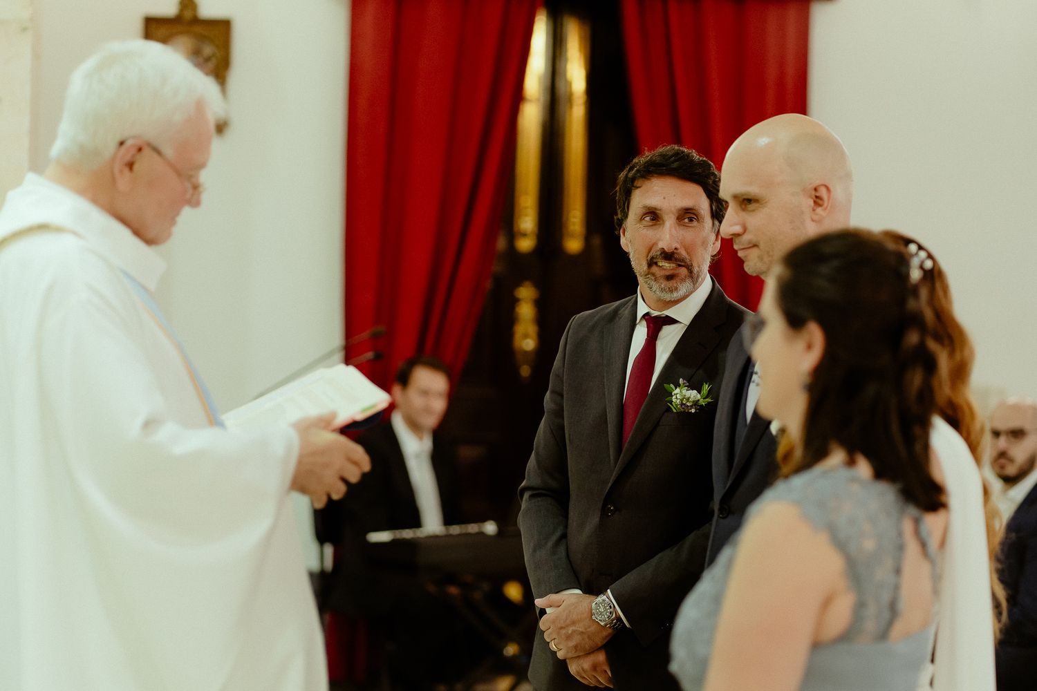 A priest officiates a wedding ceremony as the groom and best man stand before him in a church.