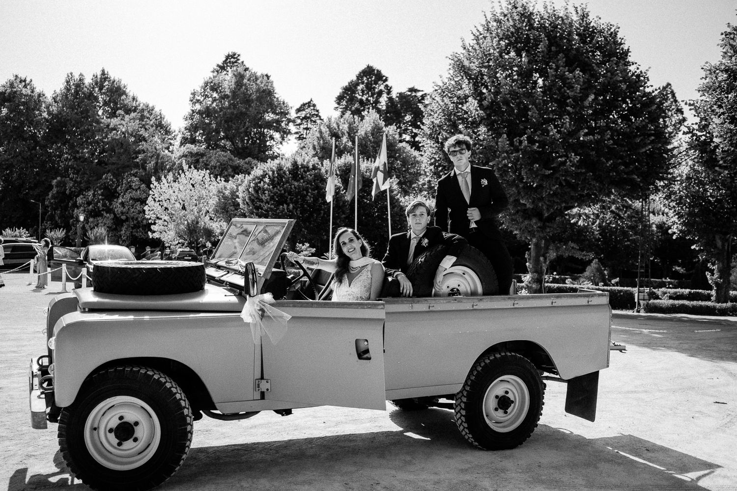Black and white photo of a wedding party riding in a classic open-top Land Rover, surrounded by trees.
