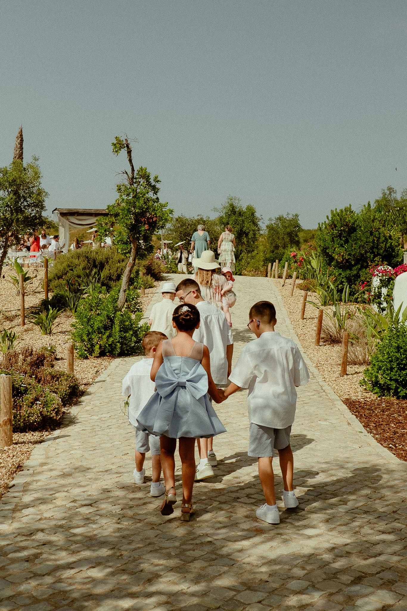 Children in white and blue outfits walk hand-in-hand along a garden path on a sunny day.
