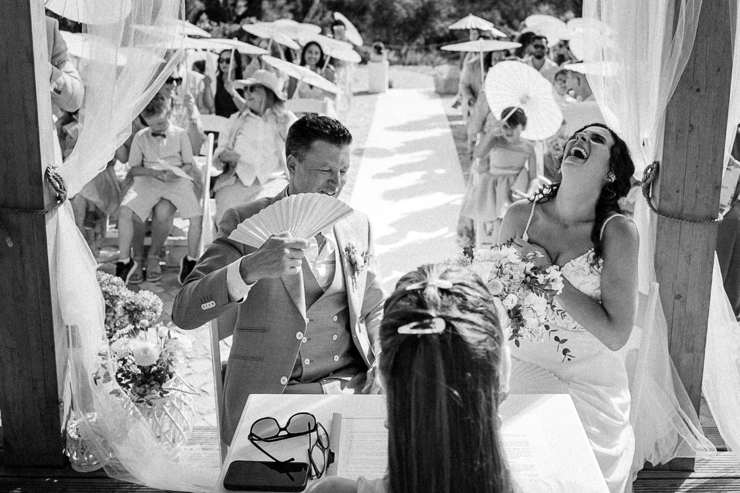 Black and white photo of a laughing bride and groom fanning themselves at an outdoor wedding ceremony.