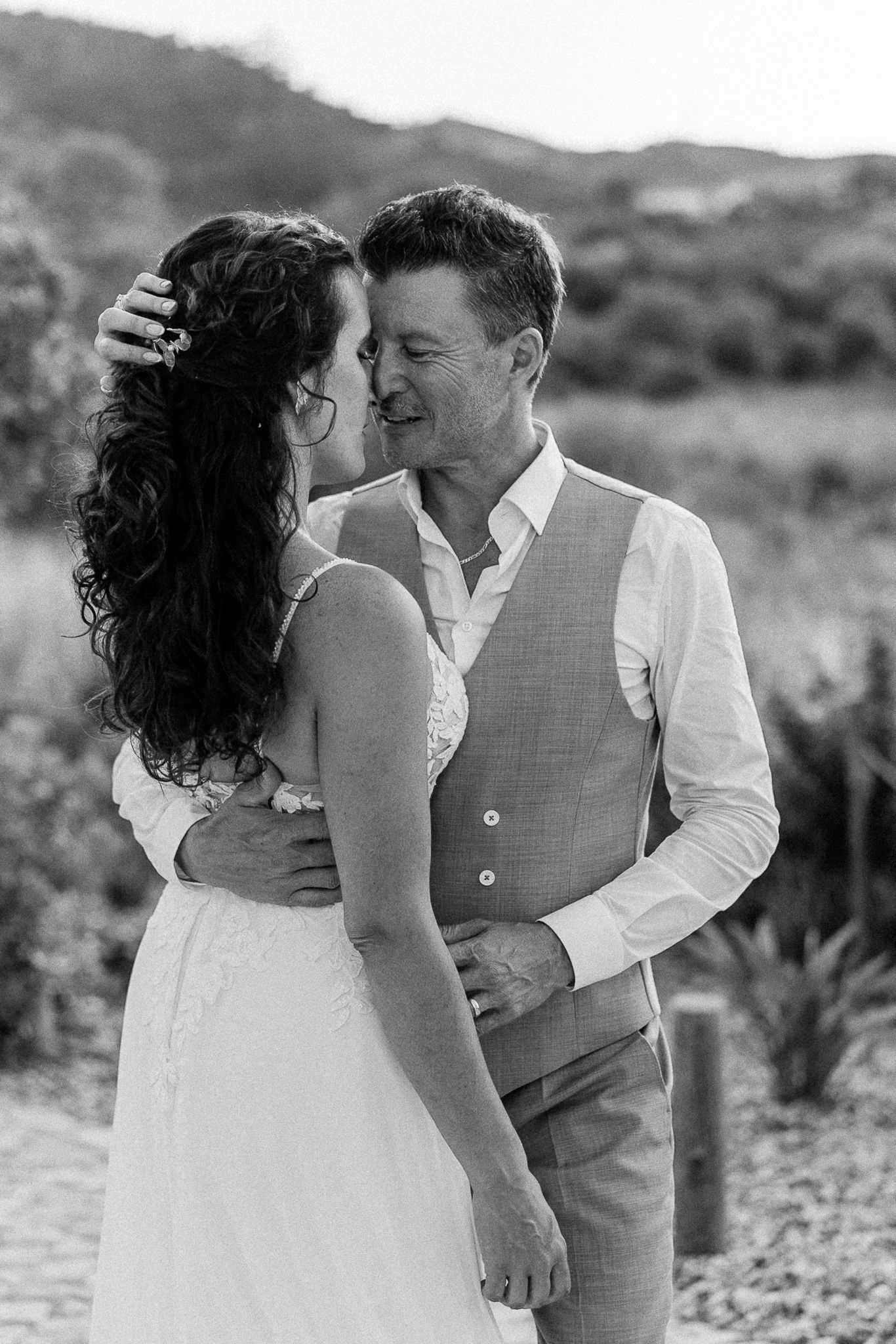Black and white photo of a couple in wedding attire sharing a romantic moment outdoors with hills in the background.