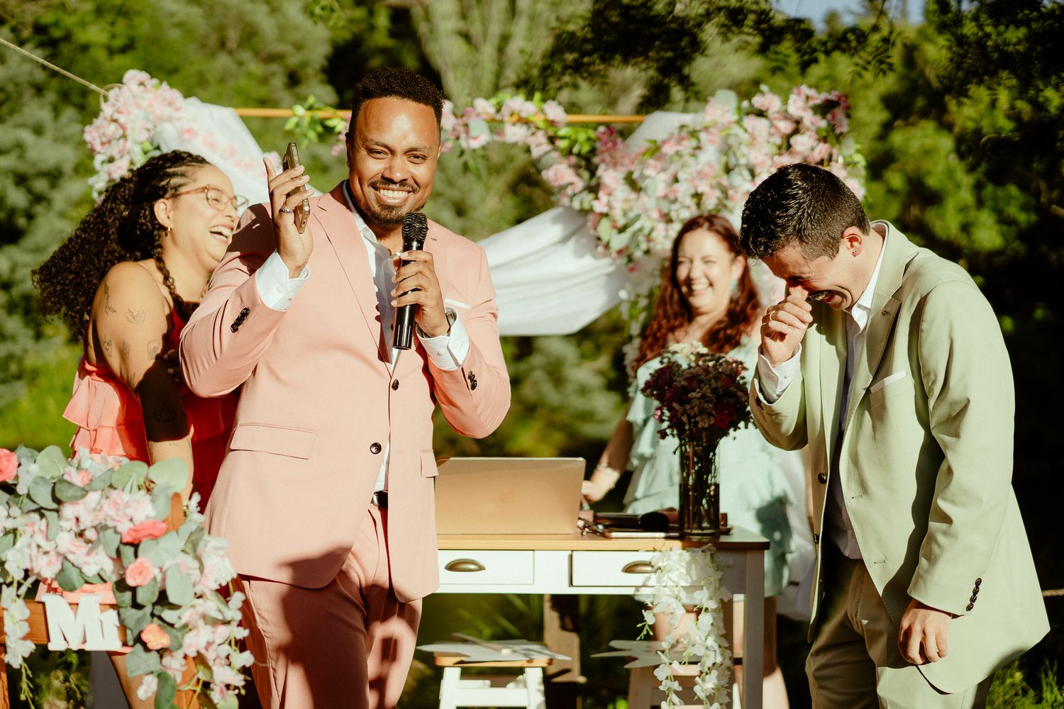 Joyful wedding ceremony outdoors with guests laughing, floral decorations, and a man in pink suit speaking at microphone.