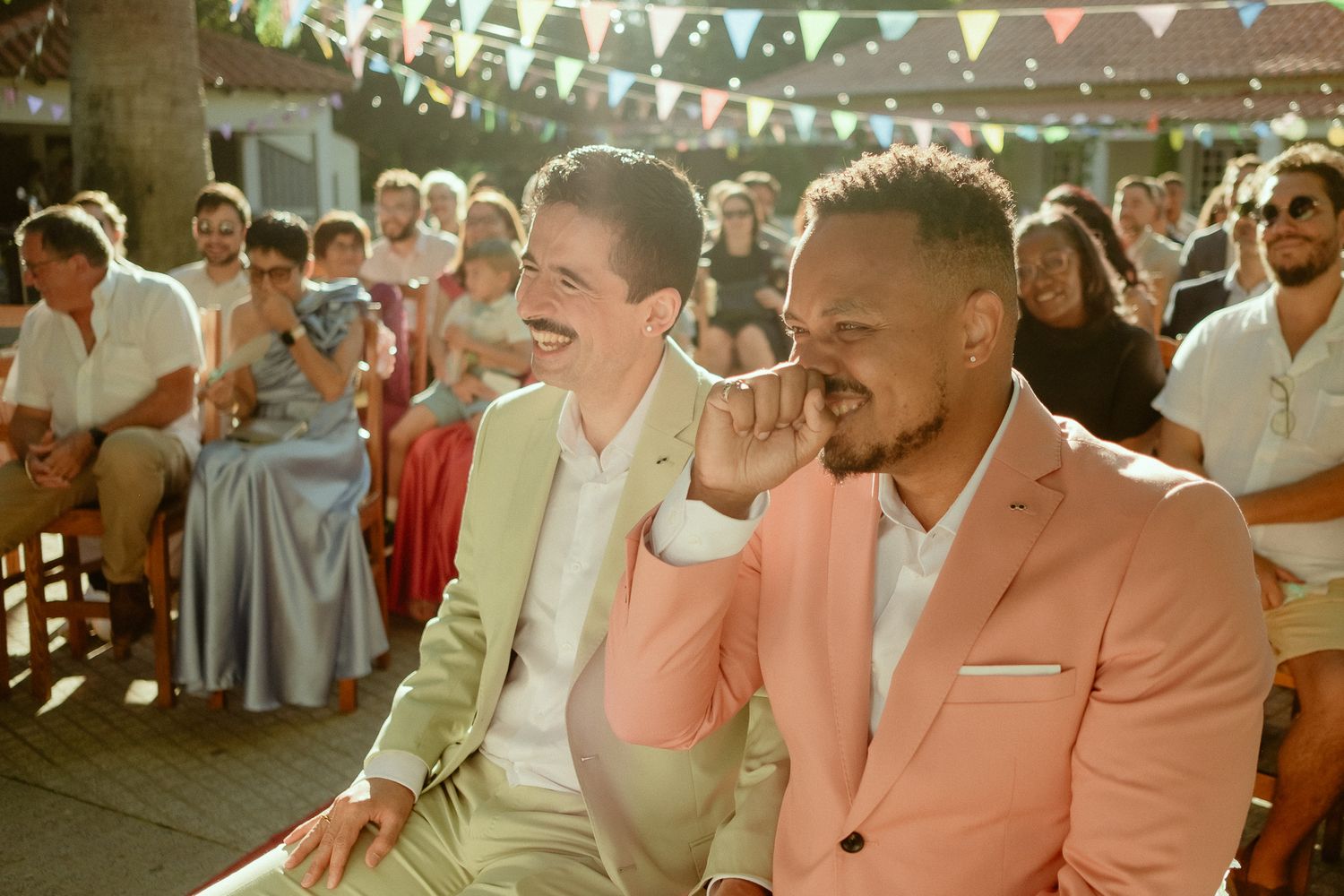 Two grooms in pink suits laugh joyfully at their outdoor wedding ceremony with guests seated behind them.