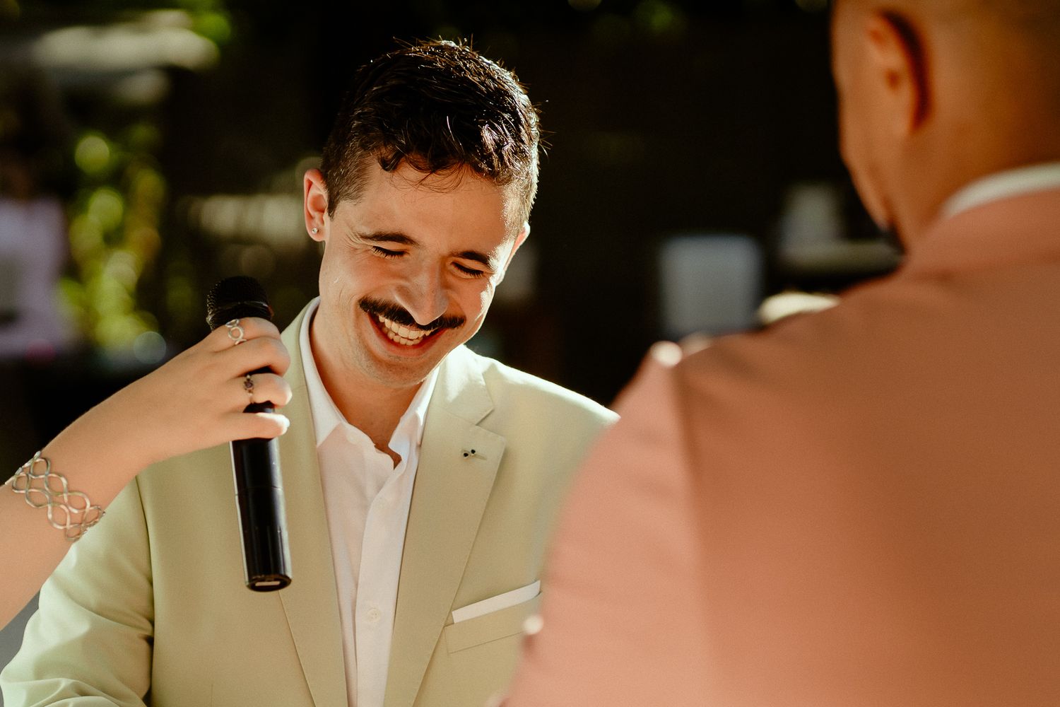 Smiling man in beige suit holding microphone at outdoor event.