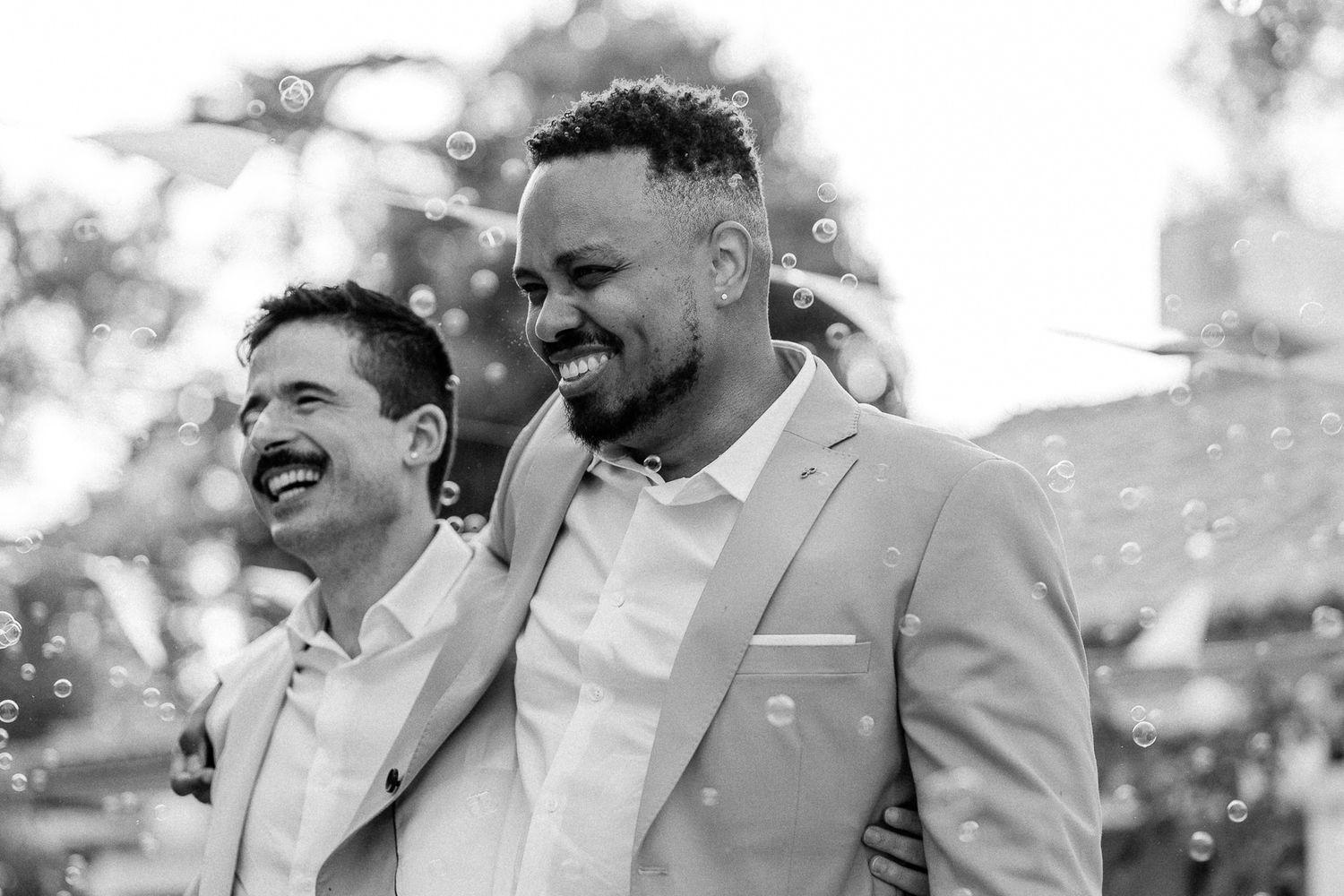 Two joyful grooms in suits laughing together outdoors surrounded by bubbles at their wedding celebration.