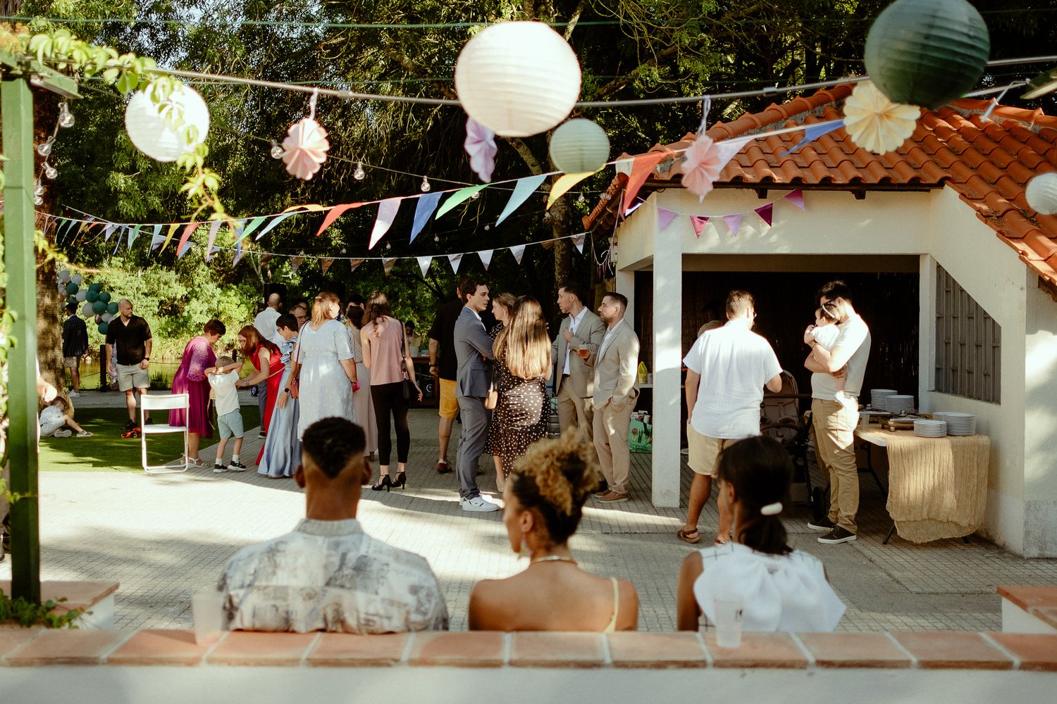 Outdoor garden party with guests mingling near a white building, decorated with colorful bunting and paper lantern lights.